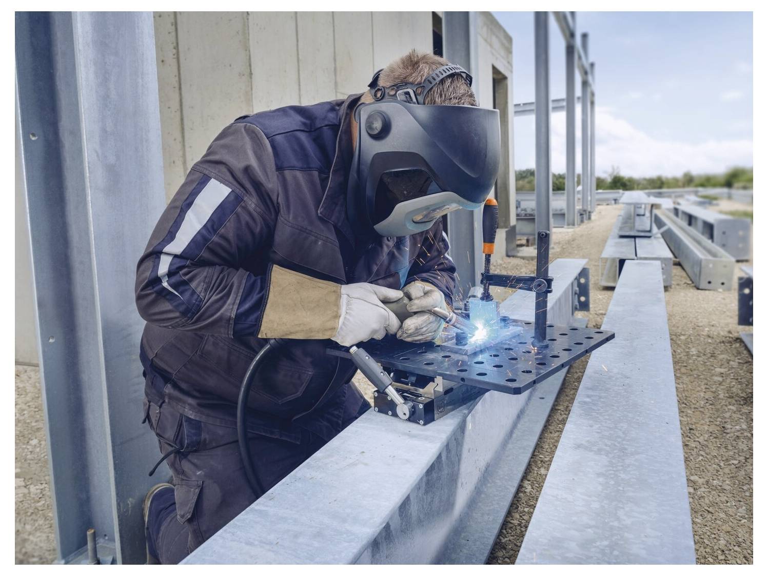 Ein Schweißer in Schutzausrüstung arbeitet an einer Metallstruktur im Freien, wobei eine helle blaue Schweißflamme sichtbar ist.
