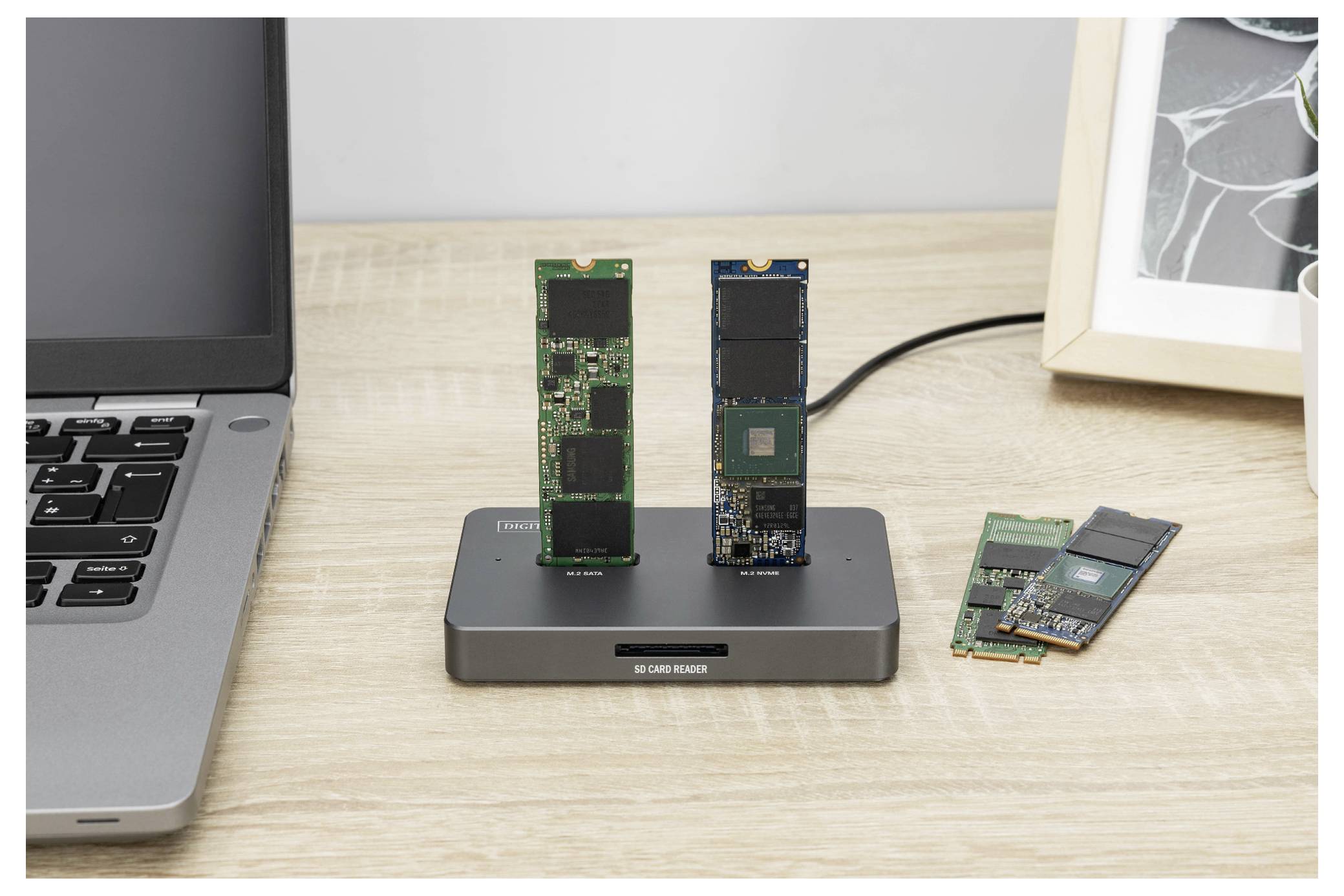 Eine Dockingstation hält zwei NVMe-SSDs aufrecht; ein Laptop befindet sich auf der linken Seite und ein gerahmtes Foto ist teilweise auf der rechten Seite sichtbar.