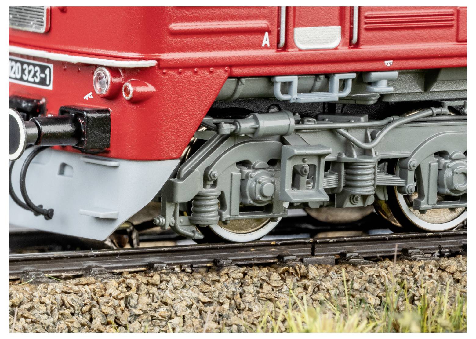 Nahaufnahme des Vorderrads und der Laufwerkskomponenten eines Modelleisenbahners auf Eisenbahnschienen, die detaillierte mechanische Komponenten und rote Außenseite zeigt.
