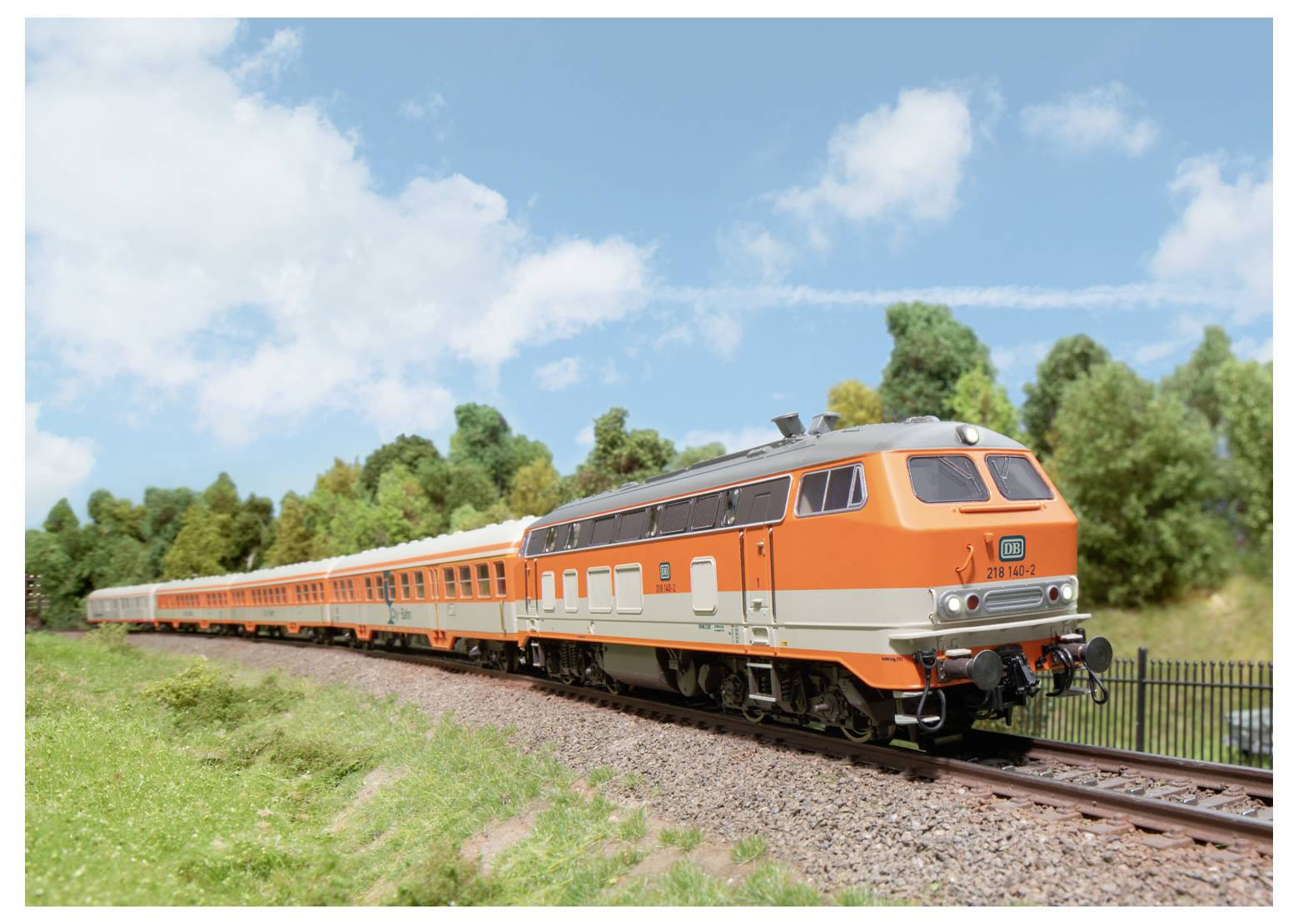 Ein hell orange- und silberfarbener Zug fährt auf einer Eisenbahnstrecke, die von üppigem Grün und Bäumen umgeben ist, unter einem teilweise bewölkten Himmel.