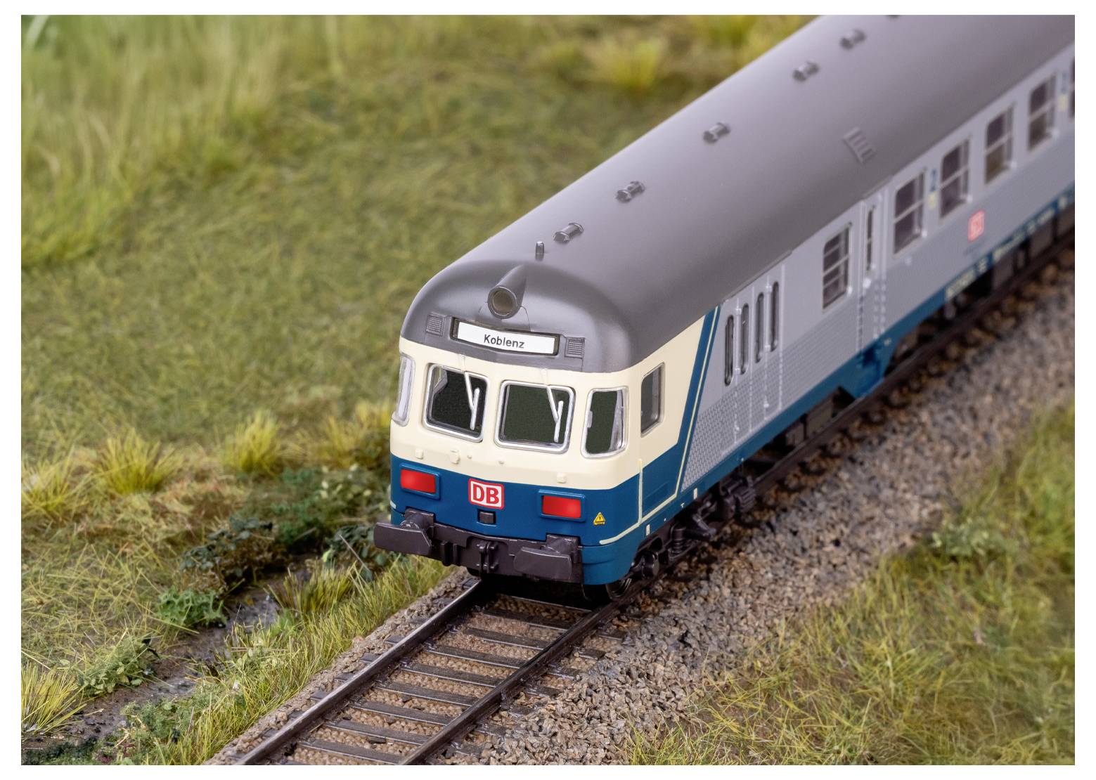 Ein orangefarbener Zug fährt durch eine üppige, grüne Landschaft mit Hügeln und Bäumen unter einem hellen, blauen Himmel mit verstreuten Wolken.