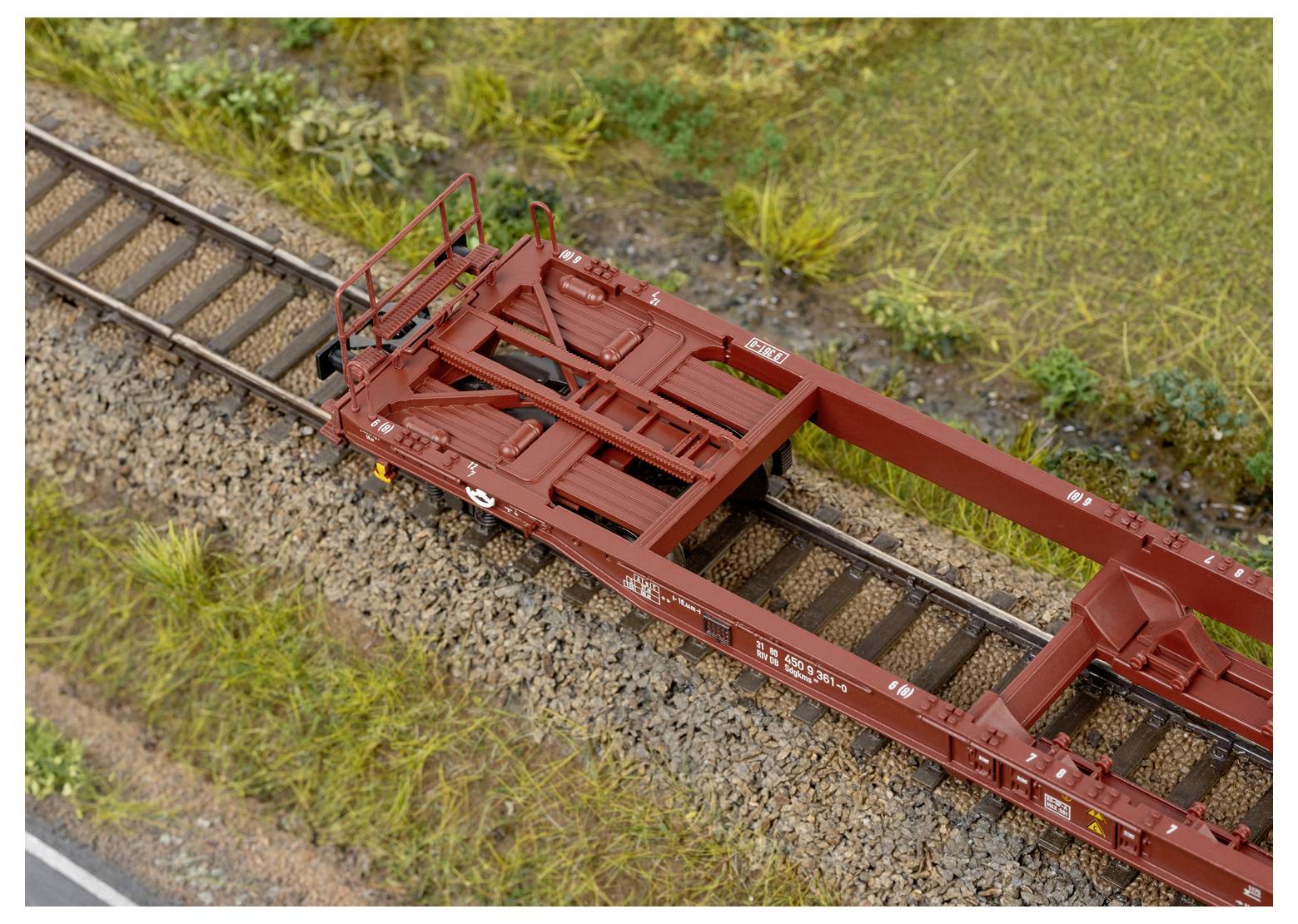 Ein Modellzug-Flachwagen mit realistischen Details sitzt auf einem Gleis, umgeben von Gras und Kies, und zeigt eine Szene aus einer Miniatureisenbahn-Anlage.