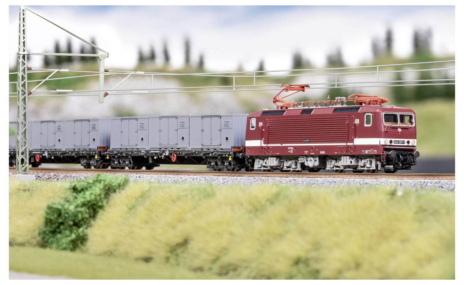 Rote elektrische Güterzuglokomotive zieht graue Güterwagen auf einer Modelleisenbahnstrecke unter Oberleitung.