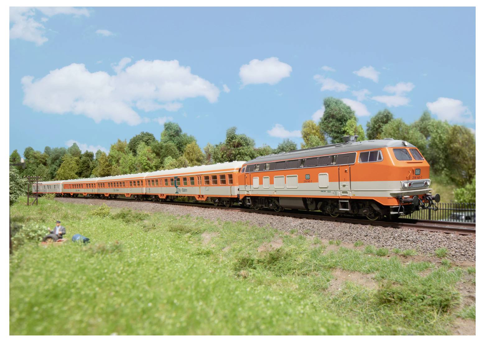 Ein orange-silberner Zug bewegt sich auf Schienen durch eine grüne Landschaft mit Bäumen und blauem Himmel, was eine malerische Reise andeutet.