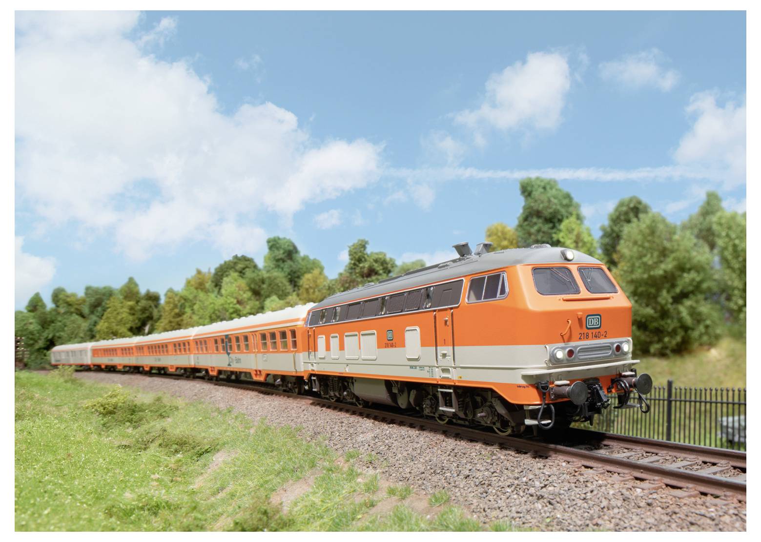 Ein Modelleisenbahn-Set mit einer orange und grau gefärbten Lokomotive zieht mehrere Personenwagen auf einem Gleis durch eine grüne Landschaft unter einem blauen Himmel.