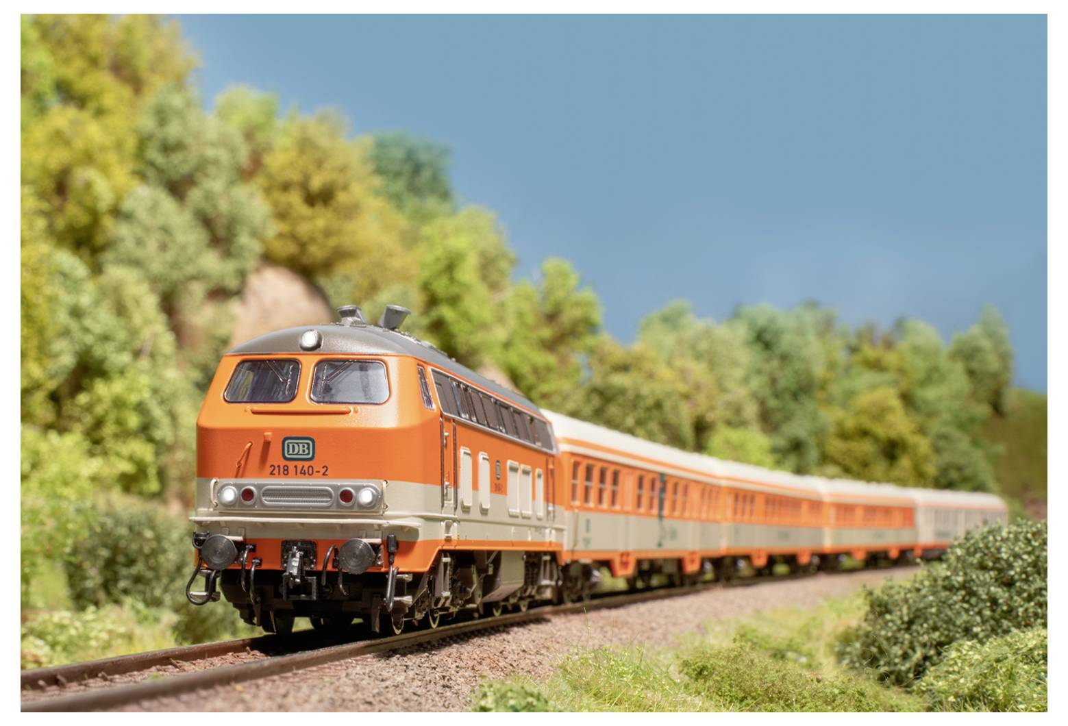Ein orangefarbener und silberner Zug fährt durch eine malerische Landschaft mit üppig grünen Bäumen unter einem klaren blauen Himmel.