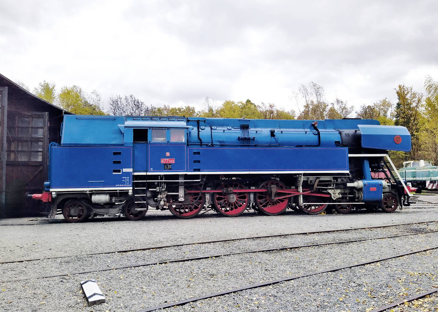 Eine große, blaue Dampflokomotive, die im Freien neben einem großen Holzgebäude ausgestellt ist, vor einem Hintergrund von Herbstbäumen und einem bewölkten Himmel.