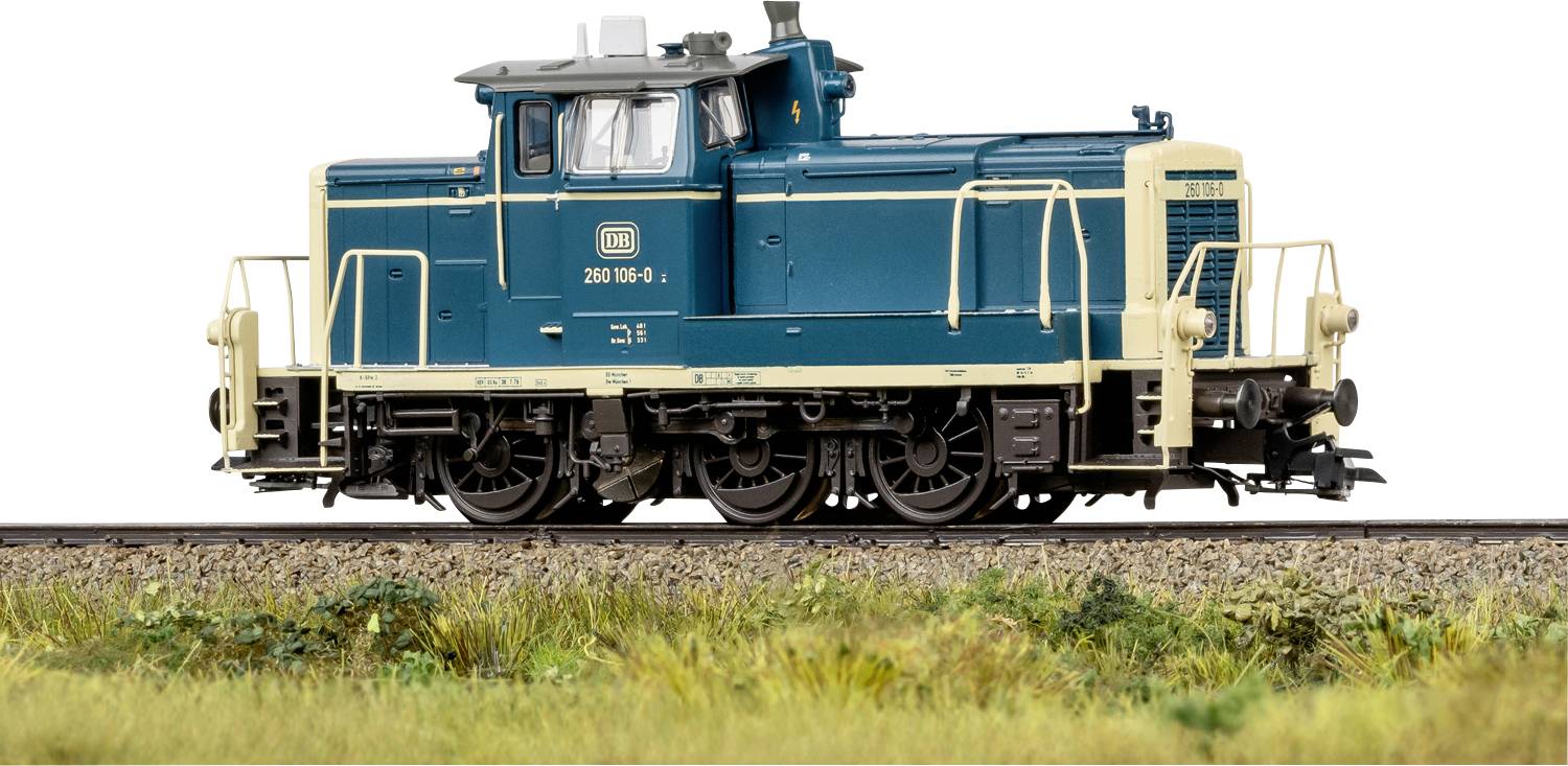 Eine blau und beige gefärbte Modelleisenbahn-Lokomotive auf einem kurzen Gleisabschnitt, vor einem weißen Hintergrund mit Gras und Kies im Vordergrund.