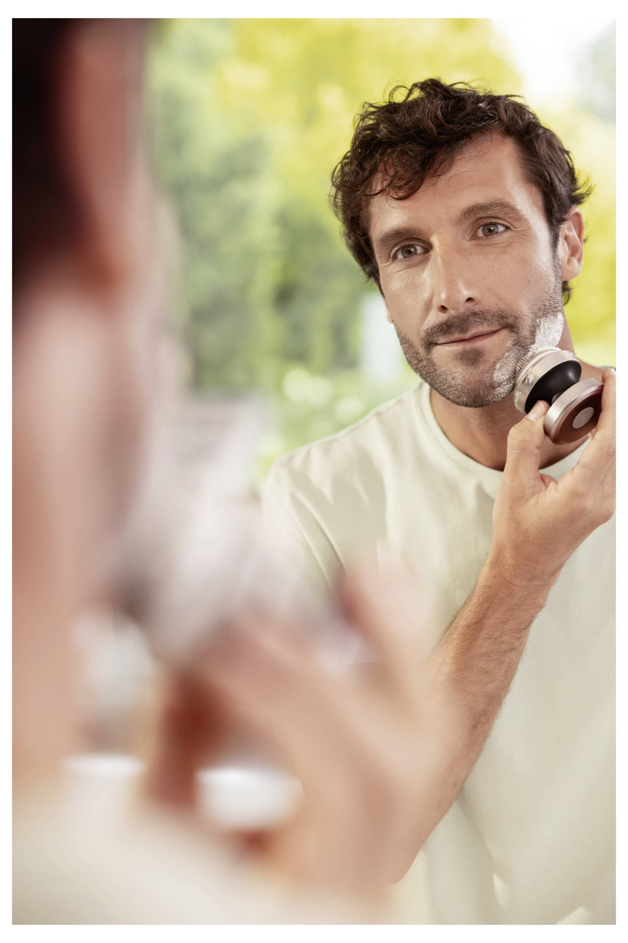 Ein Mann lächelt, während er einen Elektrorasierer vor einem Spiegel benutzt. Der Text lautet „Kompakte & handliche Form