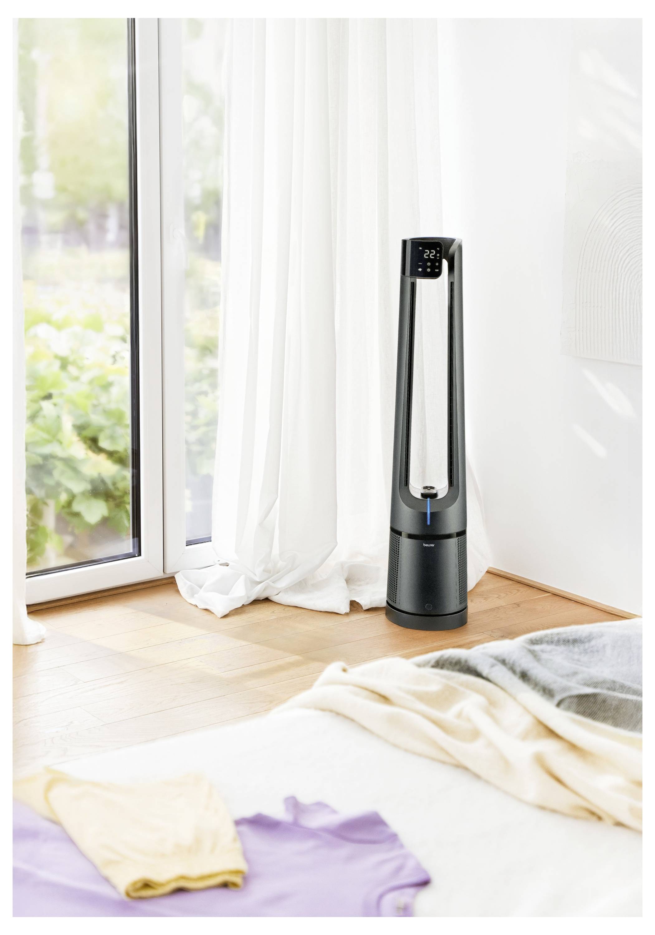 Turmventilator in modernem Zimmer neben großem Fenster, mit Sonnenlicht, das hereinfiltert, und Wäsche auf dem Bett.