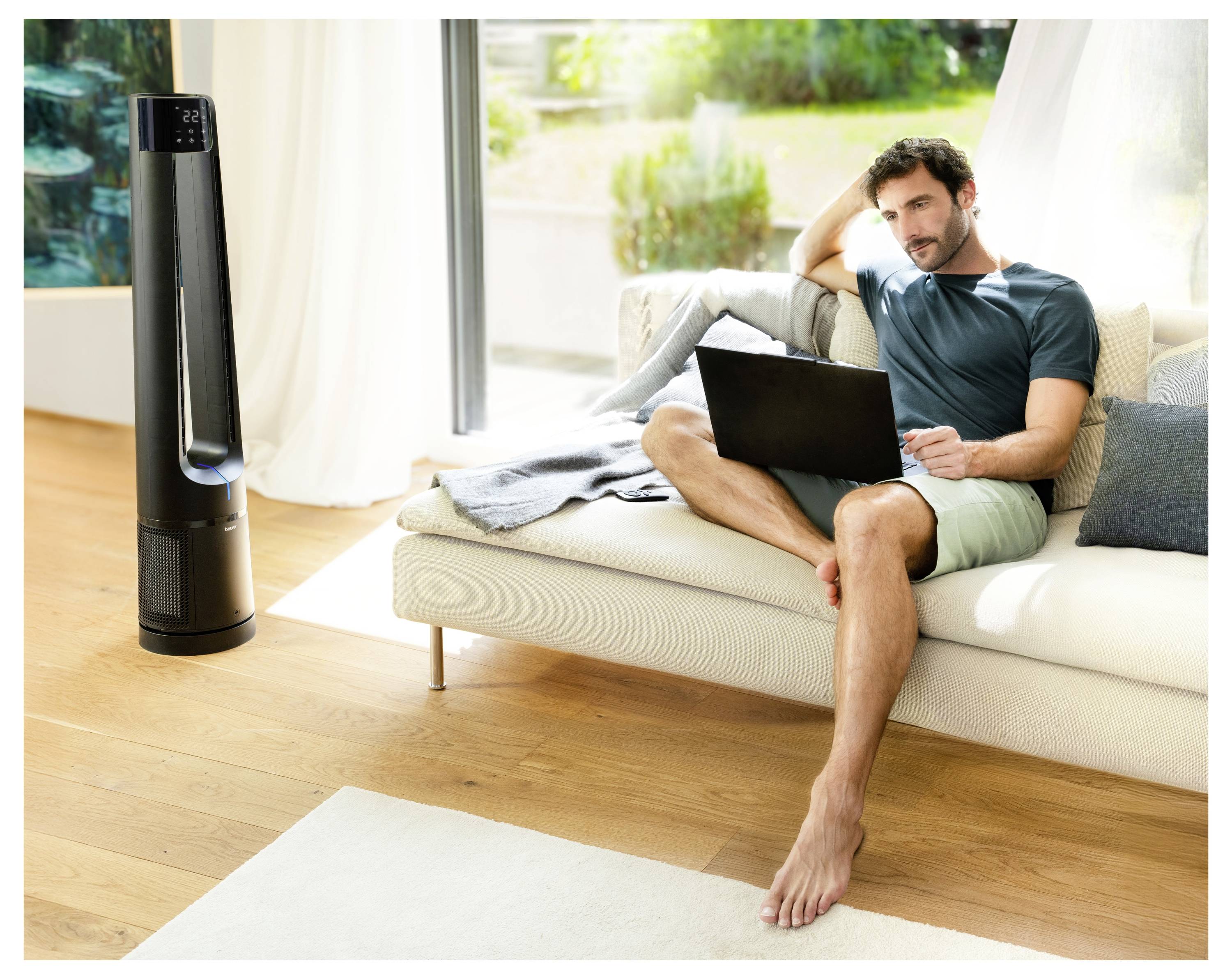 Ein Mann relaxt auf einem Sofa, benutzt einen Laptop, mit einem hohen, modernen Ventilator in der Nähe. Helles, luftiges Zimmer mit großen Fenstern und Grünflächen außerhalb.