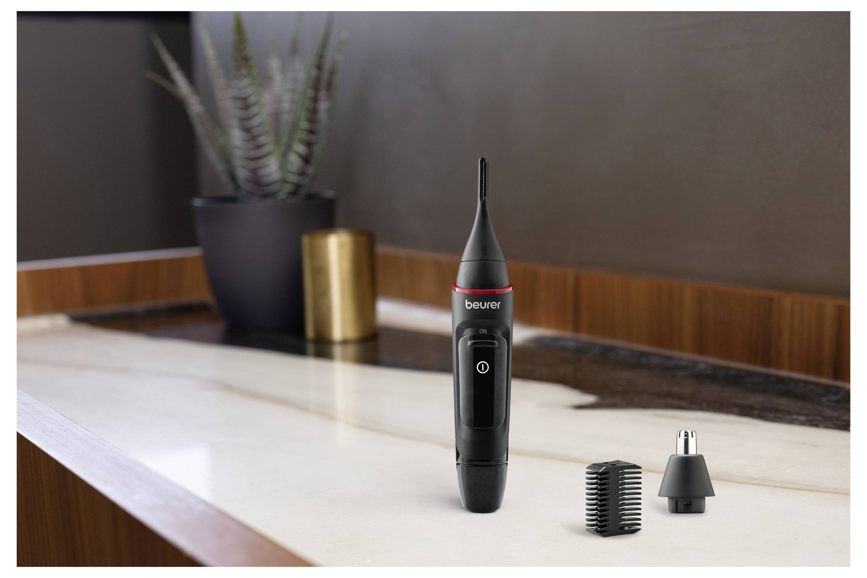 Ein schwarzer Nasen- und Ohrenhaartrimmer mit austauschbaren Aufsätzen steht auf einer weißen Marmorplatte neben einer kleinen Topfpflanze.