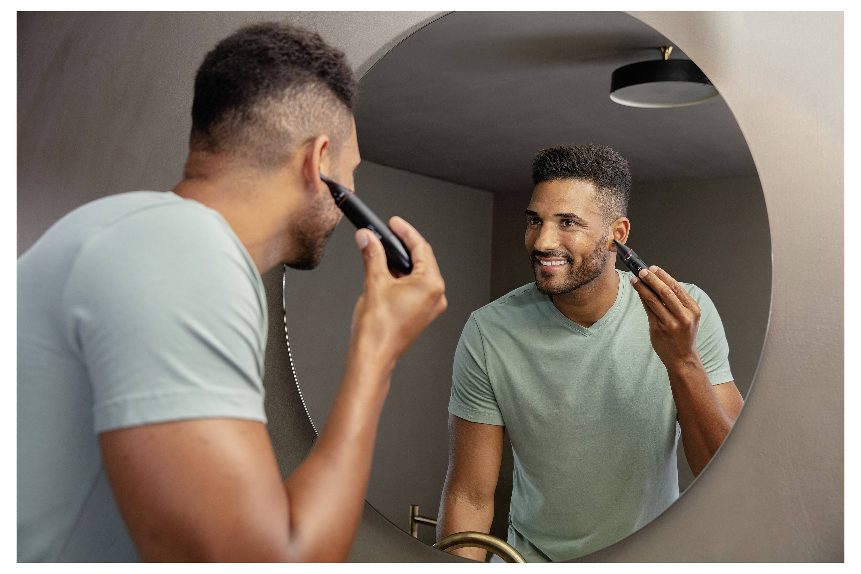 Ein Mann in einem grünen Hemd lächelt, während er sich mit einem Elektrorasierer vor einem runden Badezimmerspiegel rasiert.