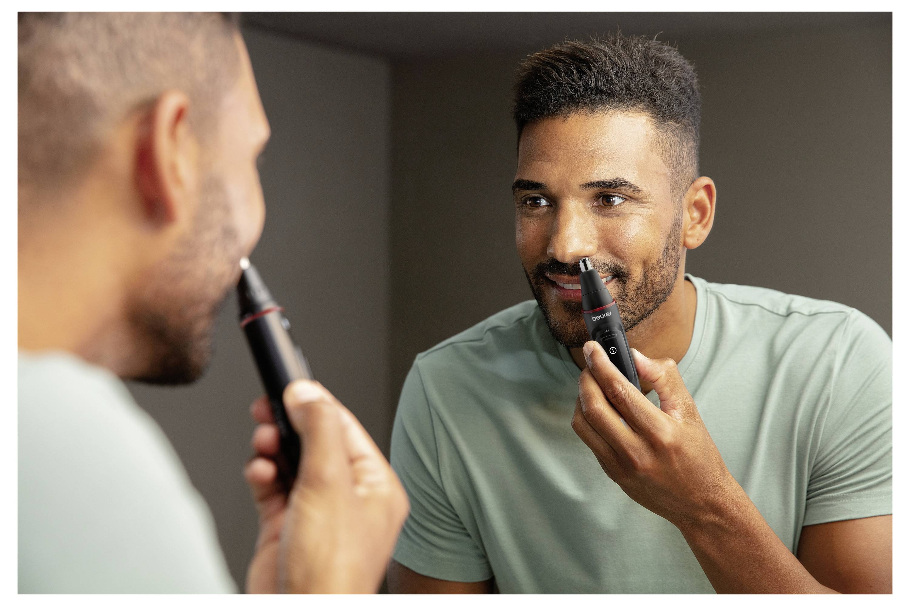 Ein Mann trimmt seinen Bart mit einem elektrischen Trimmer, während er sich im Spiegel anschaut. Er trägt ein hellgrünes T-Shirt in einer Badezimmerumgebung.