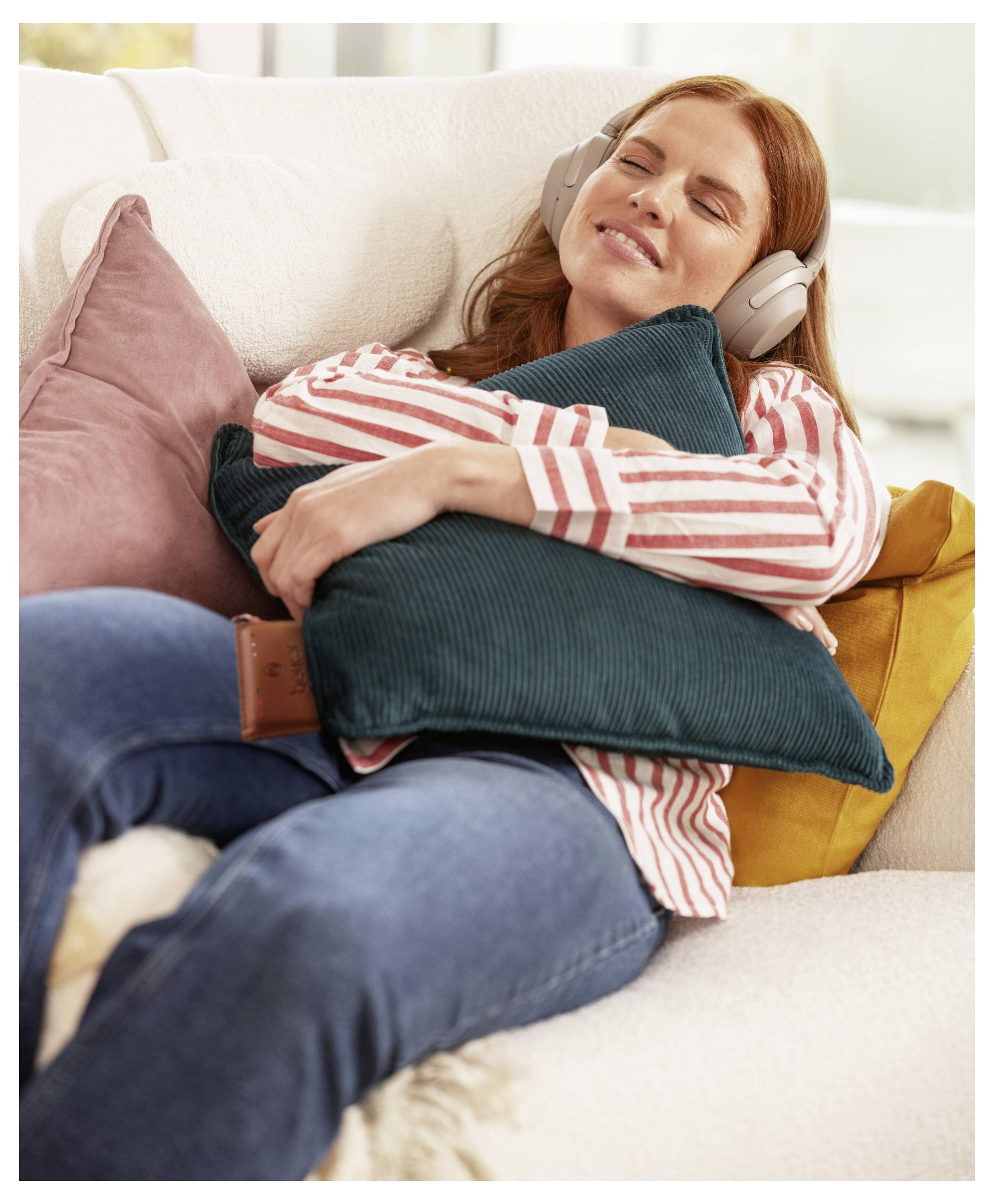 Eine Person mit Kopfhörern sitzt auf einem Sofa, hat die Augen geschlossen und lächelt, während sie ein türkises Kissen umarmt. Die Umgebung ist gemütlich und entspannt.