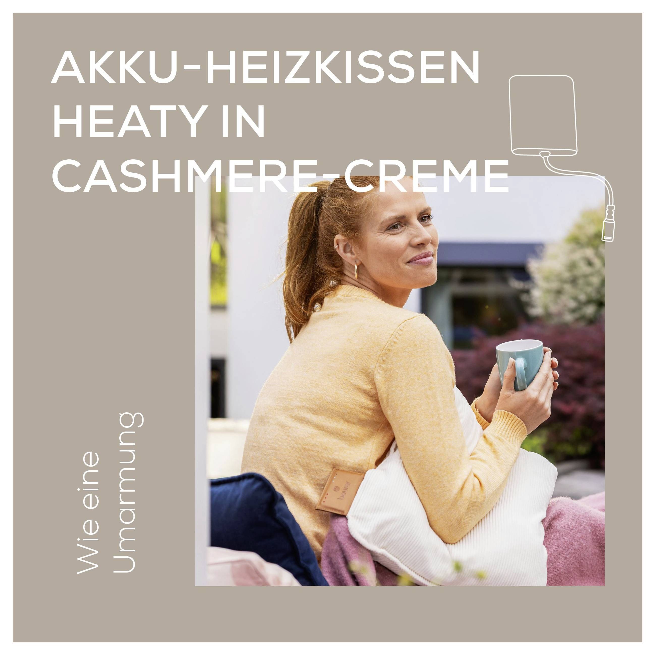 Eine Frau in einem Pfirsich-Pullover sitzt draußen auf einem Liegestuhl, hält eine blaue Tasse und ein beheiztes Kissen mit der Aufschrift „AKKU-HEIZKISSEN HEATY IN CASHMERE-CREME