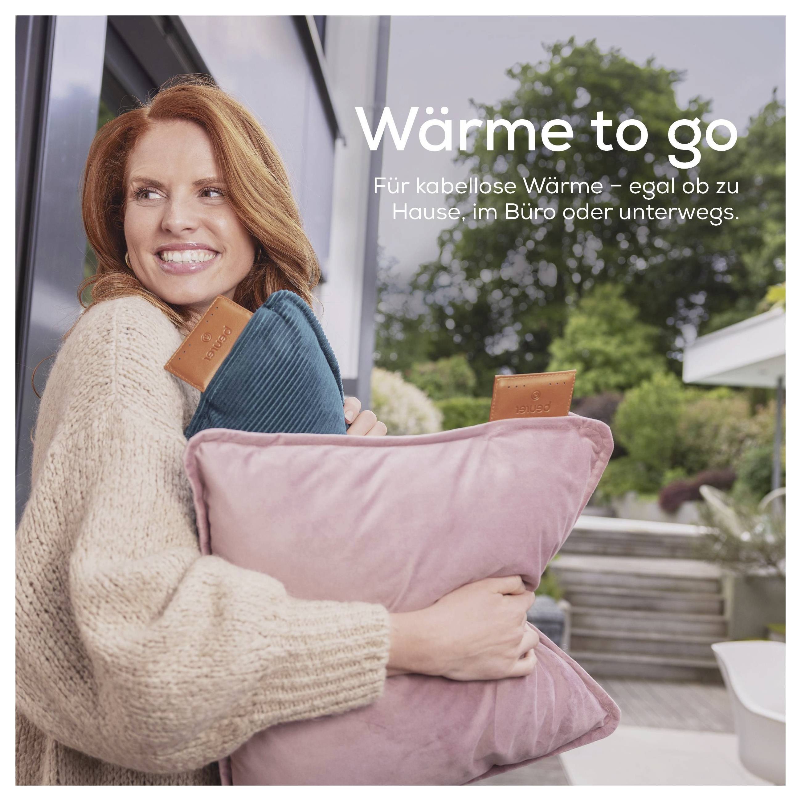 Eine Person lächelt, während sie ein flauschiges Kissen im Freien hält. Der Text lautet: „Wärme to go. Für kabellose Wärme – egal ob zu Hause, im Büro oder unterwegs.