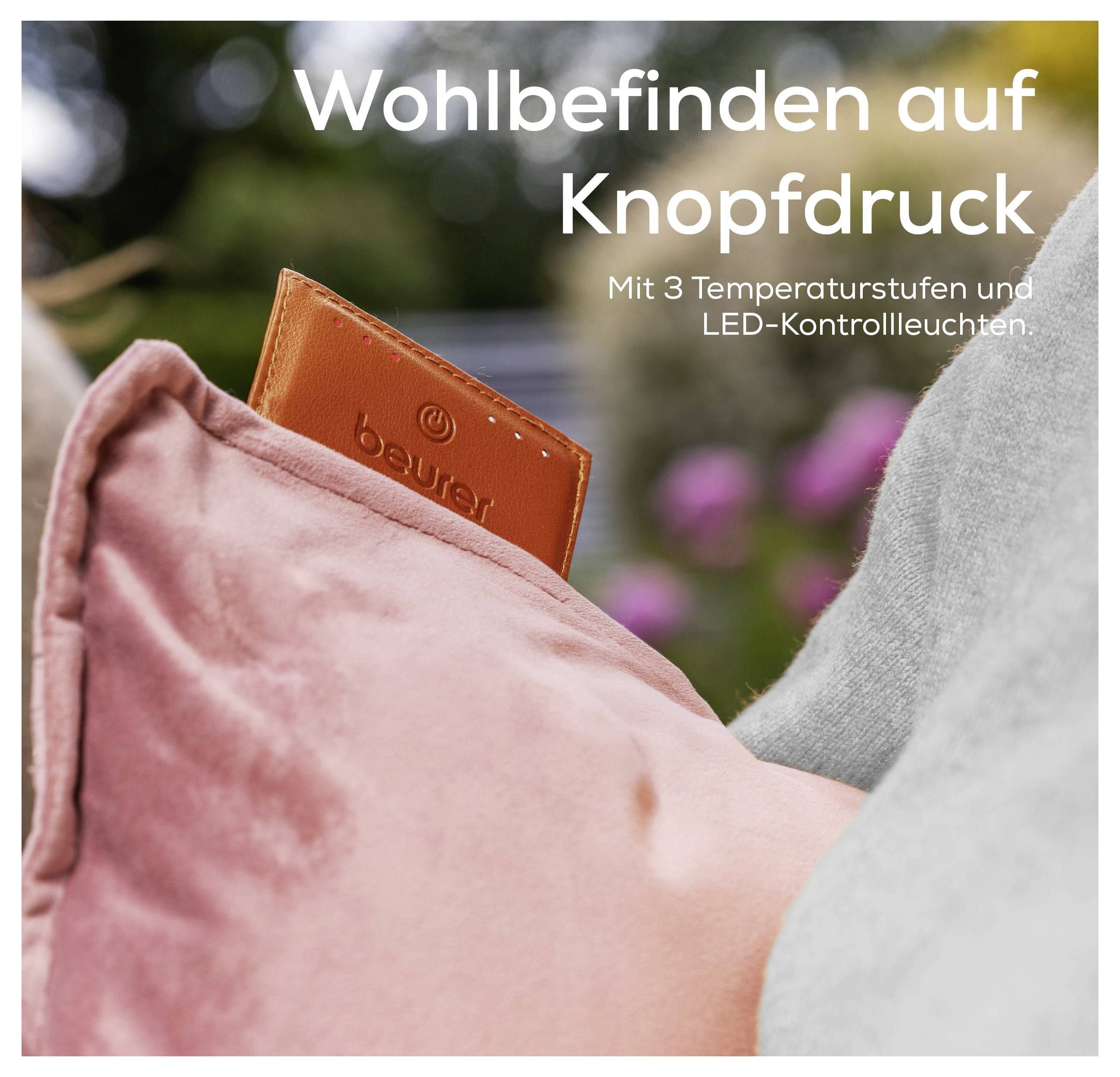 Ein braunes Beurer-Etikett taucht aus einem rosa Kissen auf und präsentiert ein Freizeitprodukt. Der Text auf Deutsch lautet: „Wohlbefinden auf Knopfdruck
