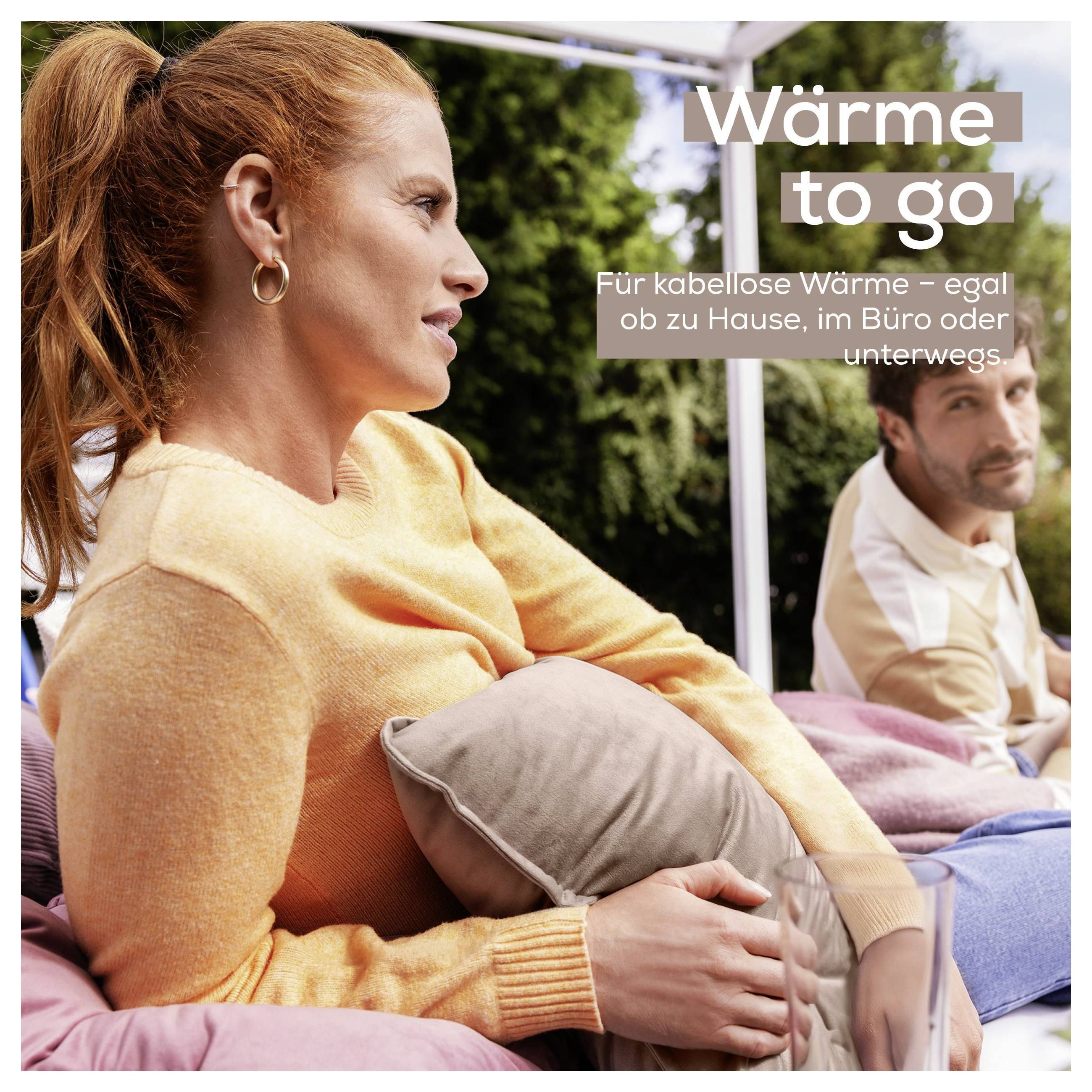 Eine Frau in einem orangefarbenen Pullover sitzt im Freien und hält ein Kissen, mit einem Mann im Hintergrund. Der Text lautet „Wärme to go