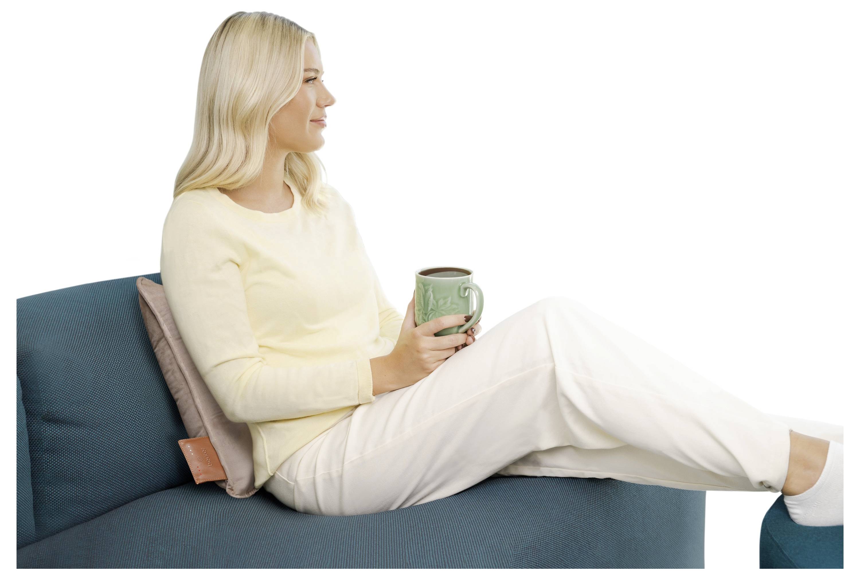 Eine Frau mit langem blondem Haar sitzt auf einem blaugrünen Sofa, trägt ein hellgelbes Oberteil und weiße Hosen, hält eine grüne Tasse in der Hand und blickt zur Seite.