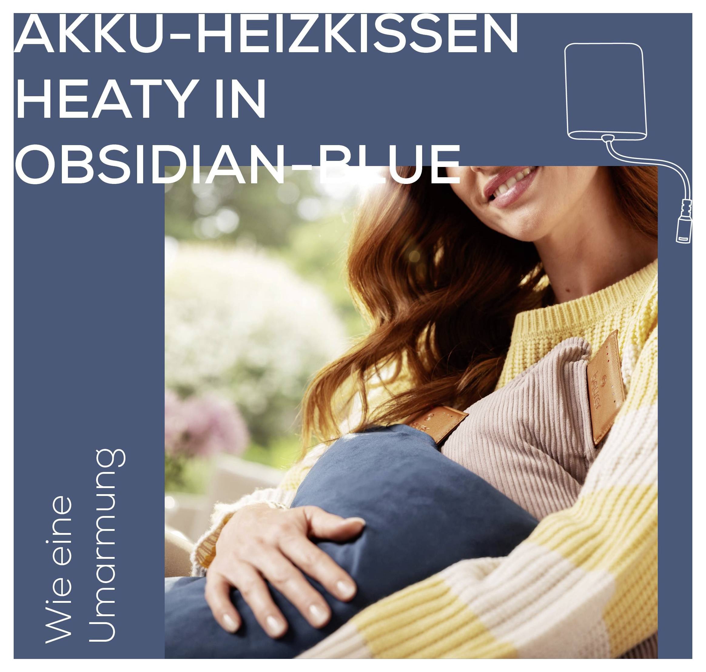 Eine Frau, lächelnd, hält ein blaues beheiztes Kissen an ihre Brust in Innenräumen. Der Text lautet „AKKU-HEIZKISSEN HEATY IN OBSIDIAN BLUE. Wie eine Umarmung.