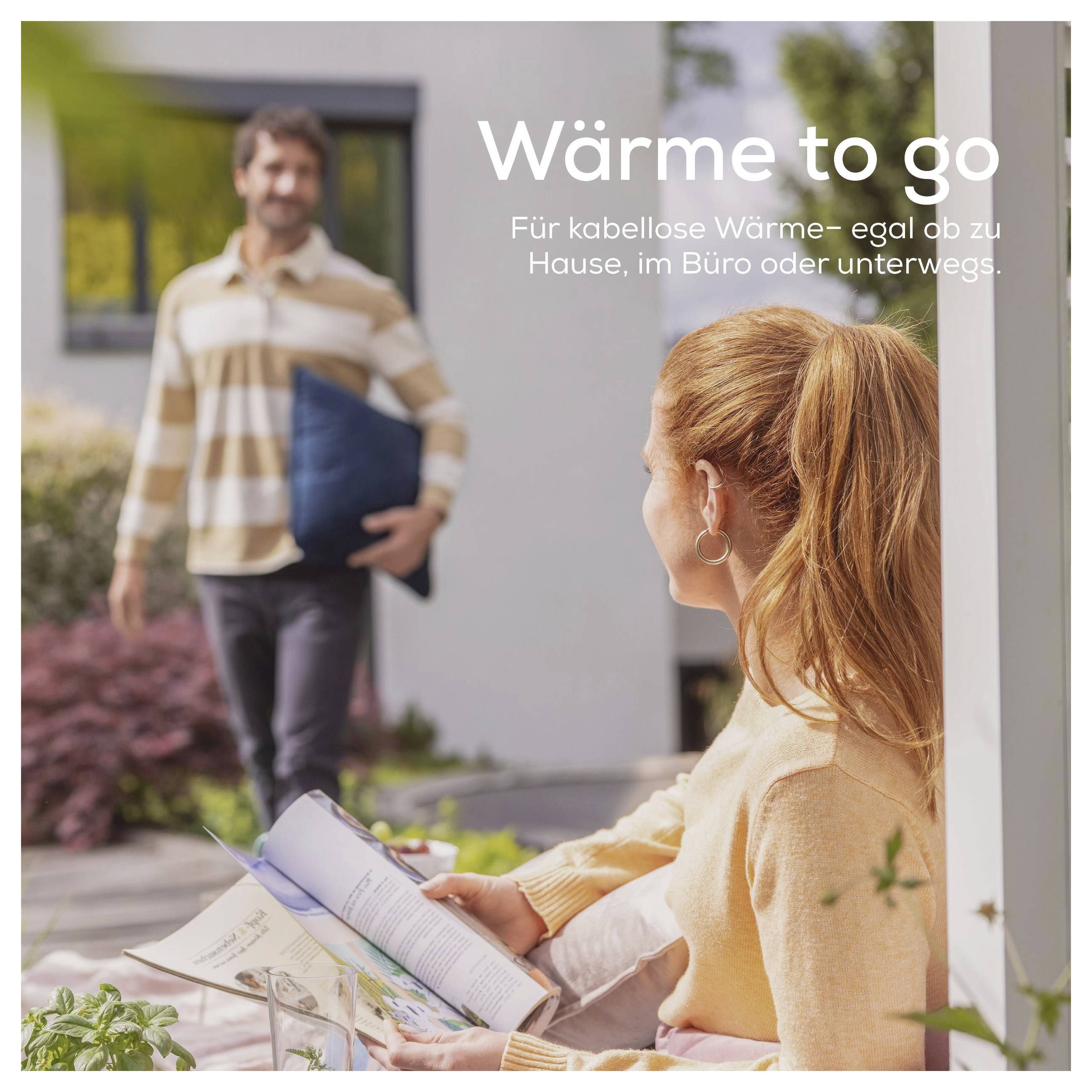 Eine Person liest eine Zeitschrift auf einer Terrasse, während sich eine andere Person mit einem Kissen nähert. Der deutsche Text bewirbt tragbare Wärme für verschiedene Umgebungen.