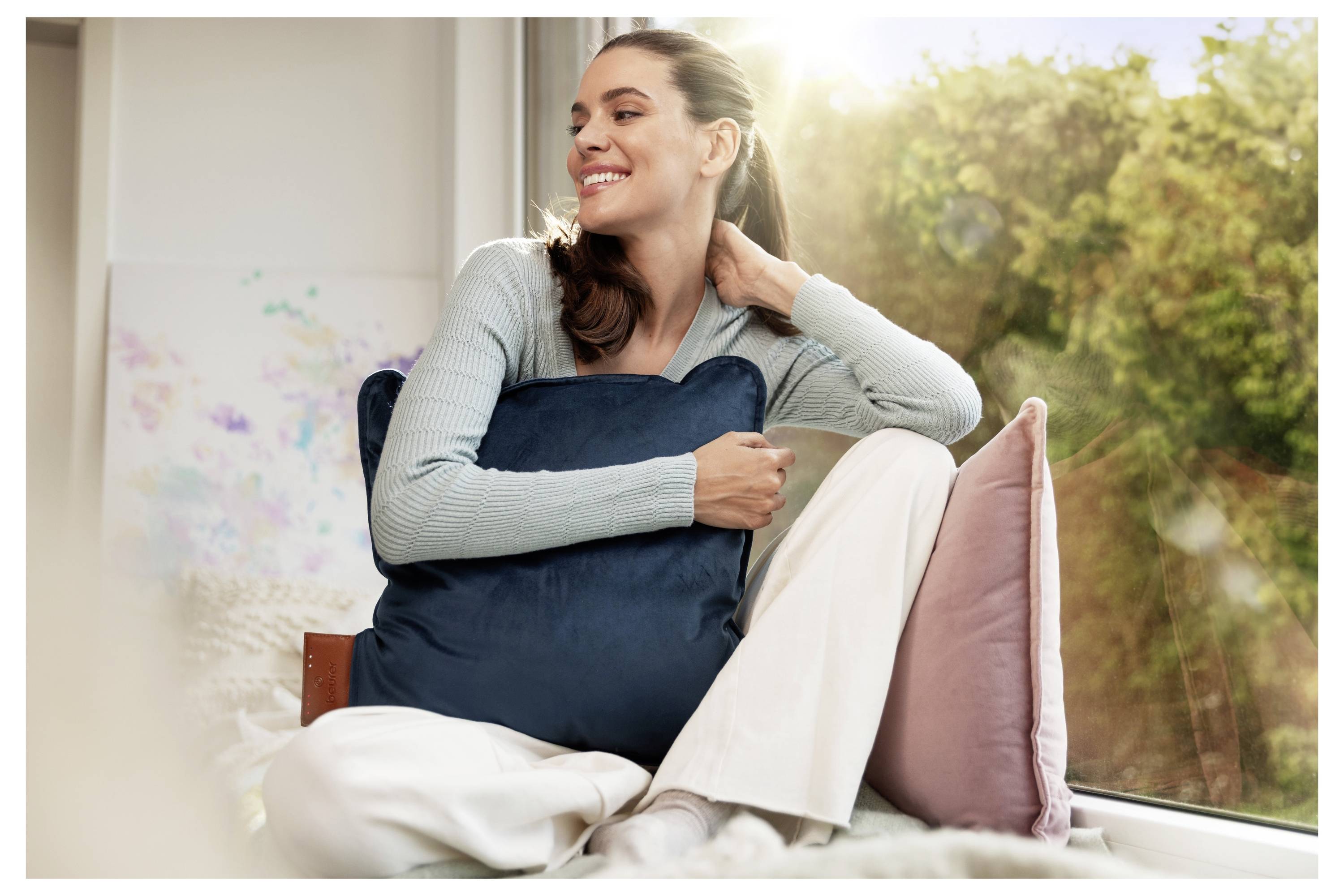 Eine Frau lächelt, während sie neben einem sonnendurchfluteten Fenster sitzt und ein dunkelblaues Kissen hält. Sie trägt einen hellblauen Pullover und weiße Hosen und genießt einen entspannten Moment.