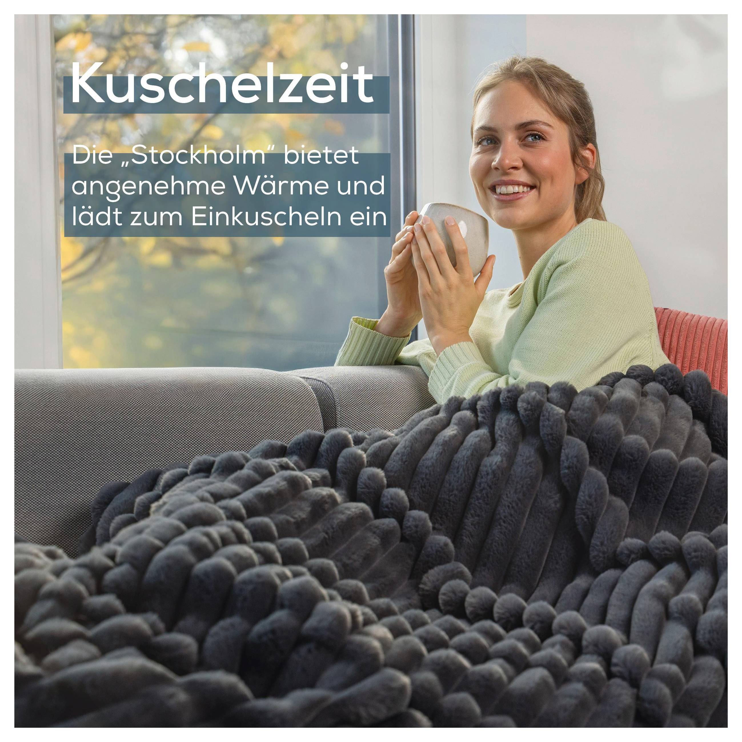 Eine Person sitzt in einer gemütlichen Umgebung auf einem Sofa, eingewickelt in eine dicke, strukturierte Decke und hält eine Tasse, mit großen Fenstern im Hintergrund.