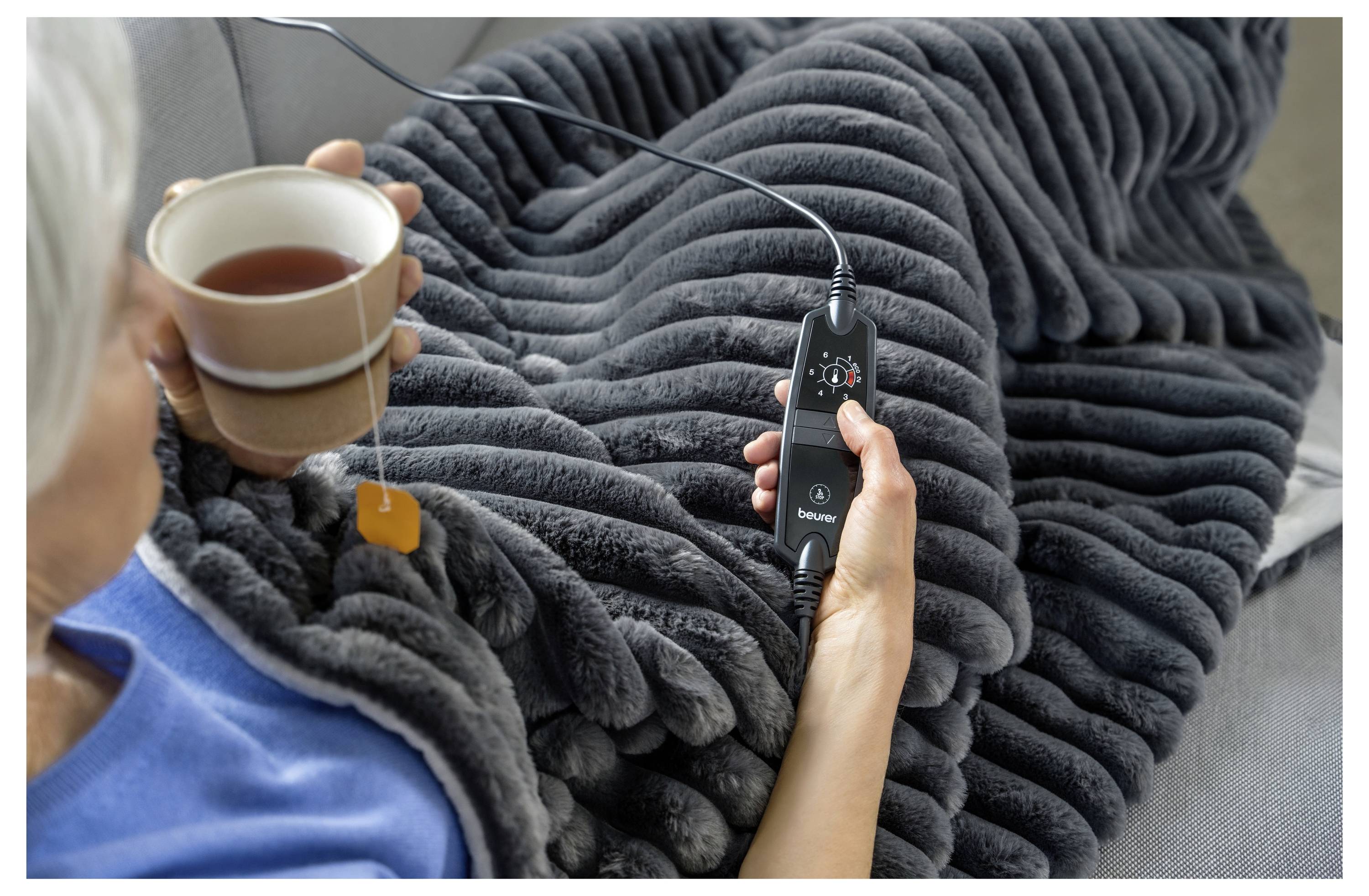 Person in eine kuschelige graue Decke eingewickelt, hält ein heißes Getränk und eine Fernbedienung, möglicherweise um die Temperatureinstellungen der Decke anzupassen.