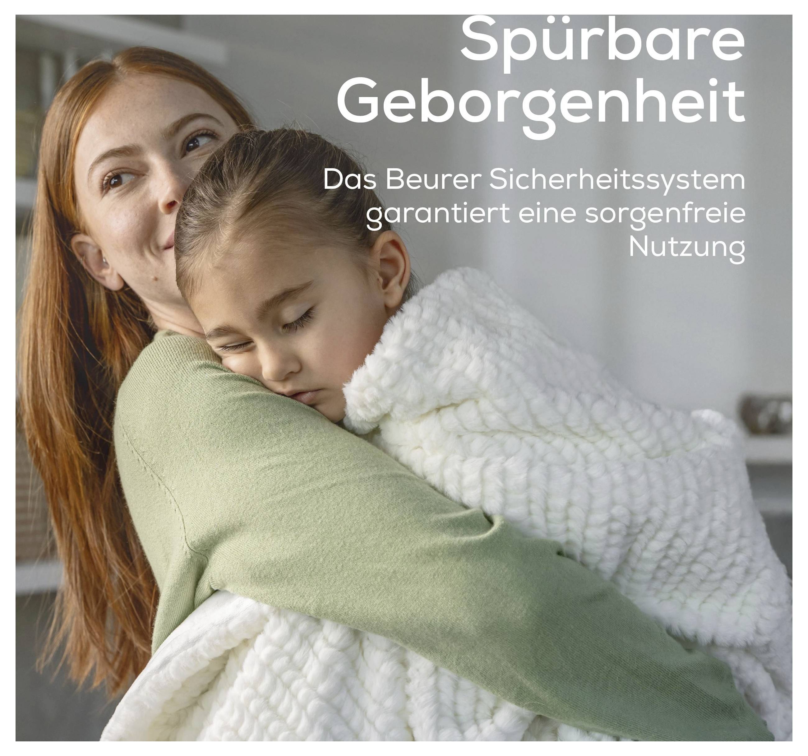 Eine Frau lächelt, während sie ein schlafendes Kind hält, das in eine weiße Decke gewickelt ist. Der deutsche Text lautet: „Greifbare Sicherheit. Das Beurer-Sicherheitssystem garantiert sorgenfreie Nutzung.