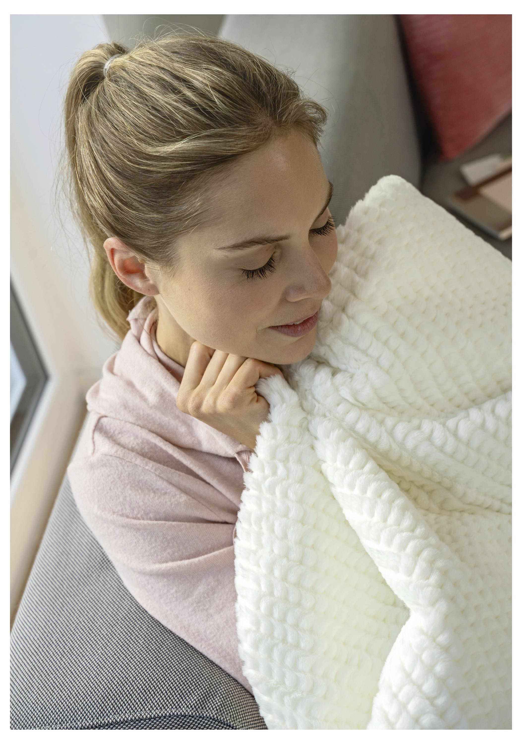 Eine Person hält sanft eine strukturierte weiße Decke, während sie drinnen sitzt, trägt ein rosa Oberteil und hat einen ruhigen Gesichtsausdruck.