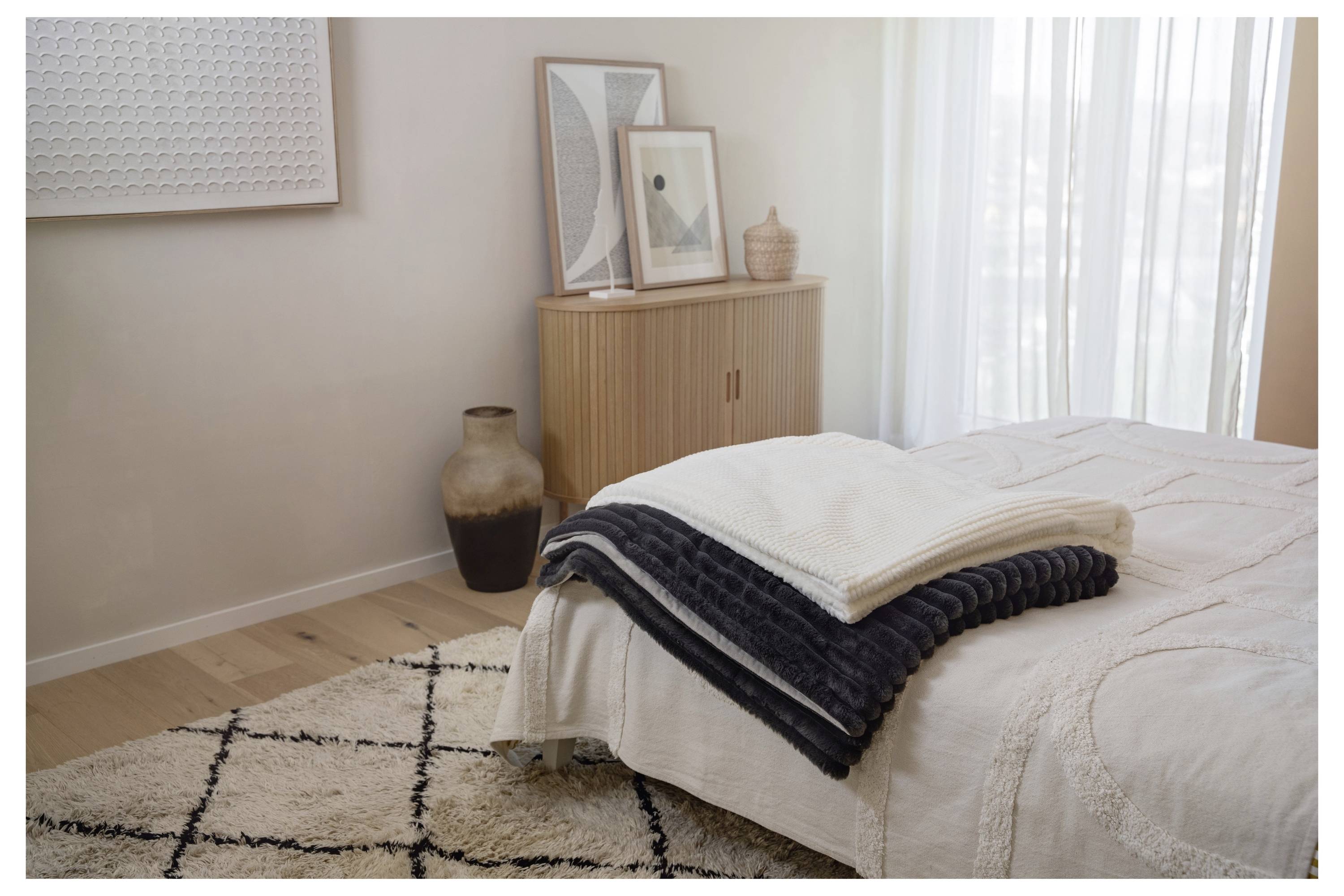 Ein ordentlich gemachtes Bett mit grauen und weißen Decken, ein gemusterter Teppich auf Holzboden, ein Holzschrank mit gerahmter Kunst und eine große Vase in der Nähe.