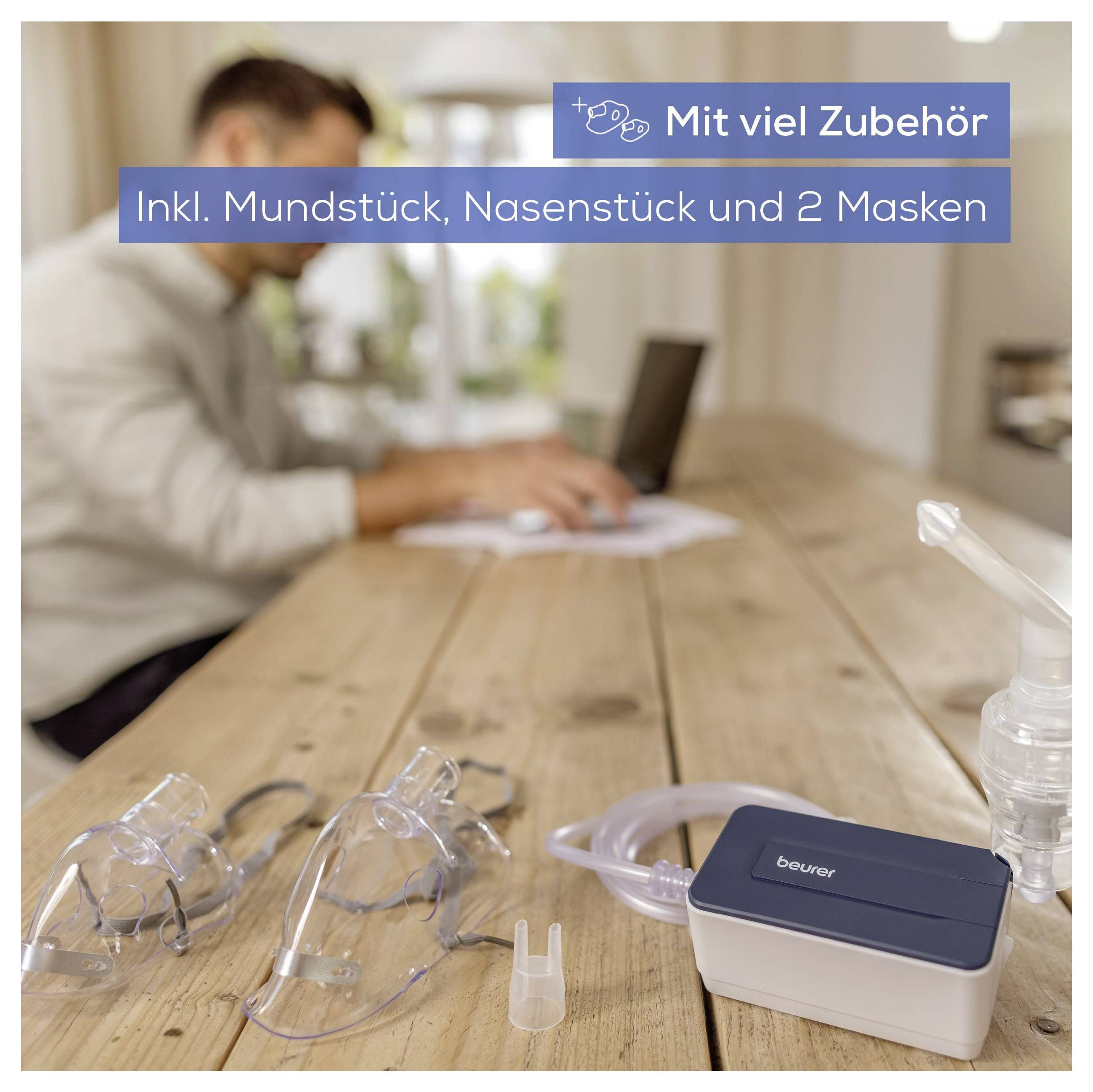 Ein Zerstäuber-Set auf einem Tisch umfasst ein Mundstück, ein Nasenstück und zwei Masken. Eine Person arbeitet im Hintergrund an einem Laptop.