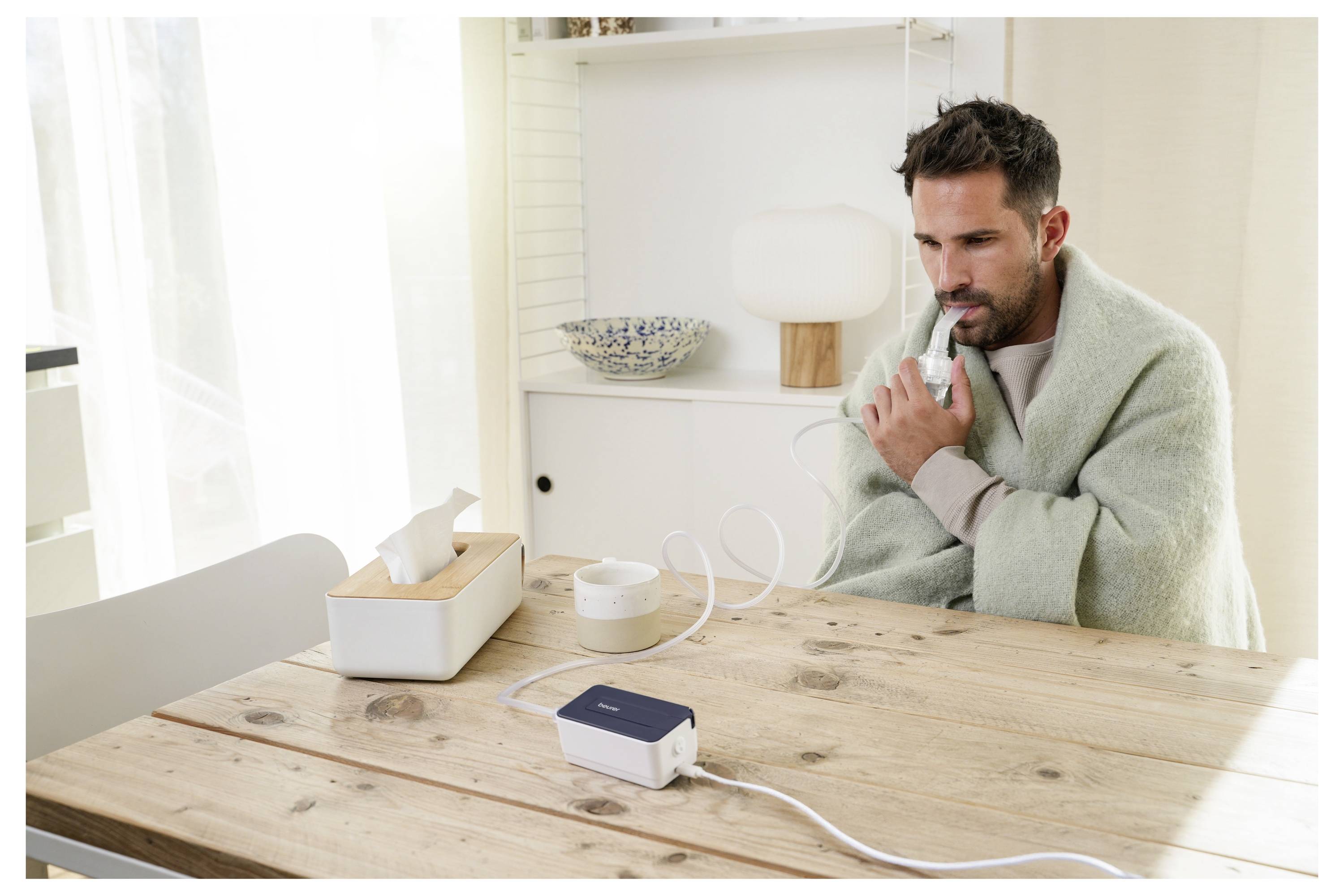 Ein Mann sitzt an einem Holztisch und benutzt einen Zerstäuber, in eine Decke gehüllt. Eine Taschentuchbox und eine kleine Schüssel befinden sich in der Nähe auf dem Tisch.