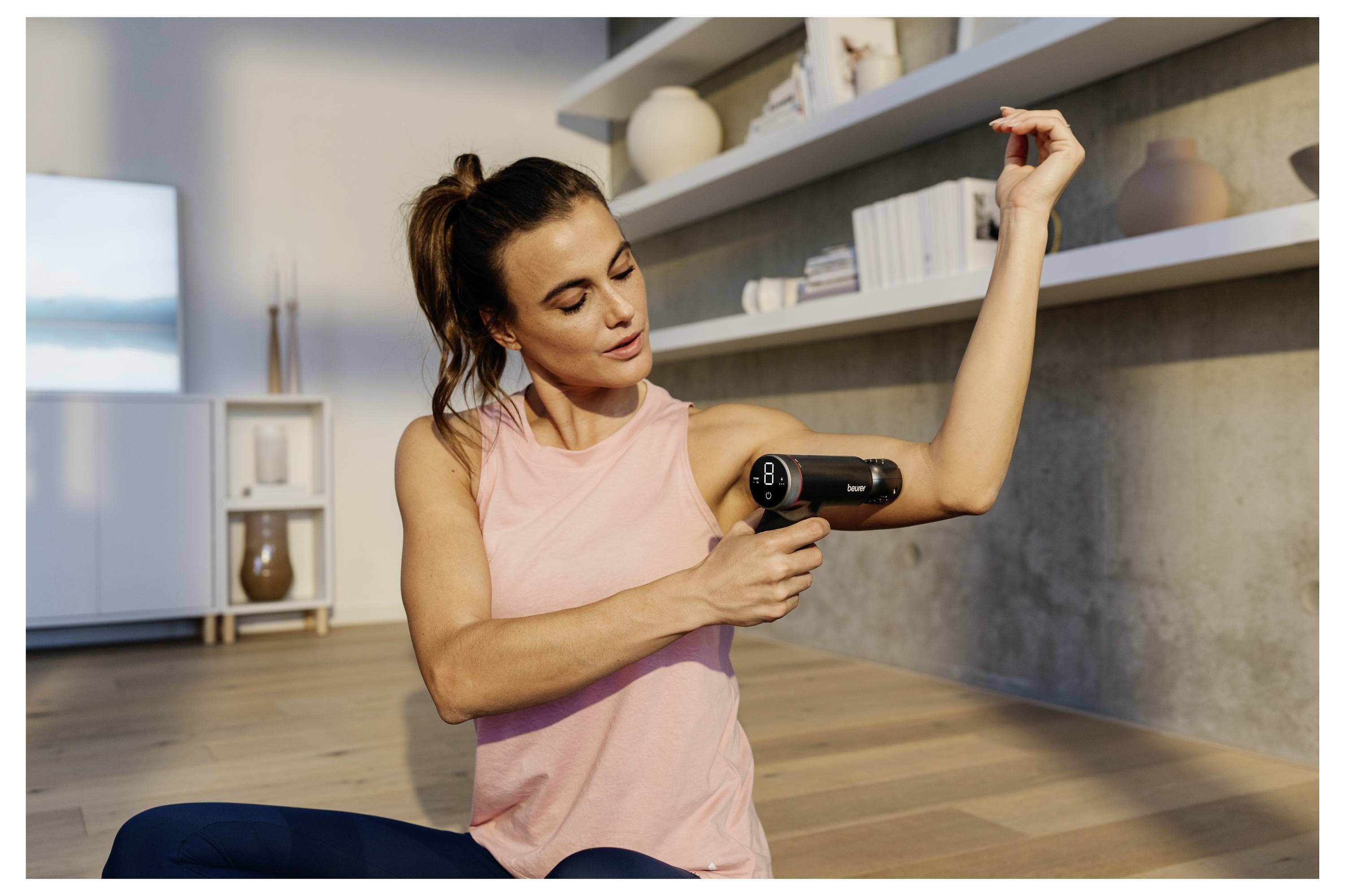 Eine Frau benutzt eine Massagepistole an ihrem Arm, während sie auf dem Boden in einem modernen, hellen Raum sitzt. Im Hintergrund sind Regale mit Dekorationselementen sichtbar.