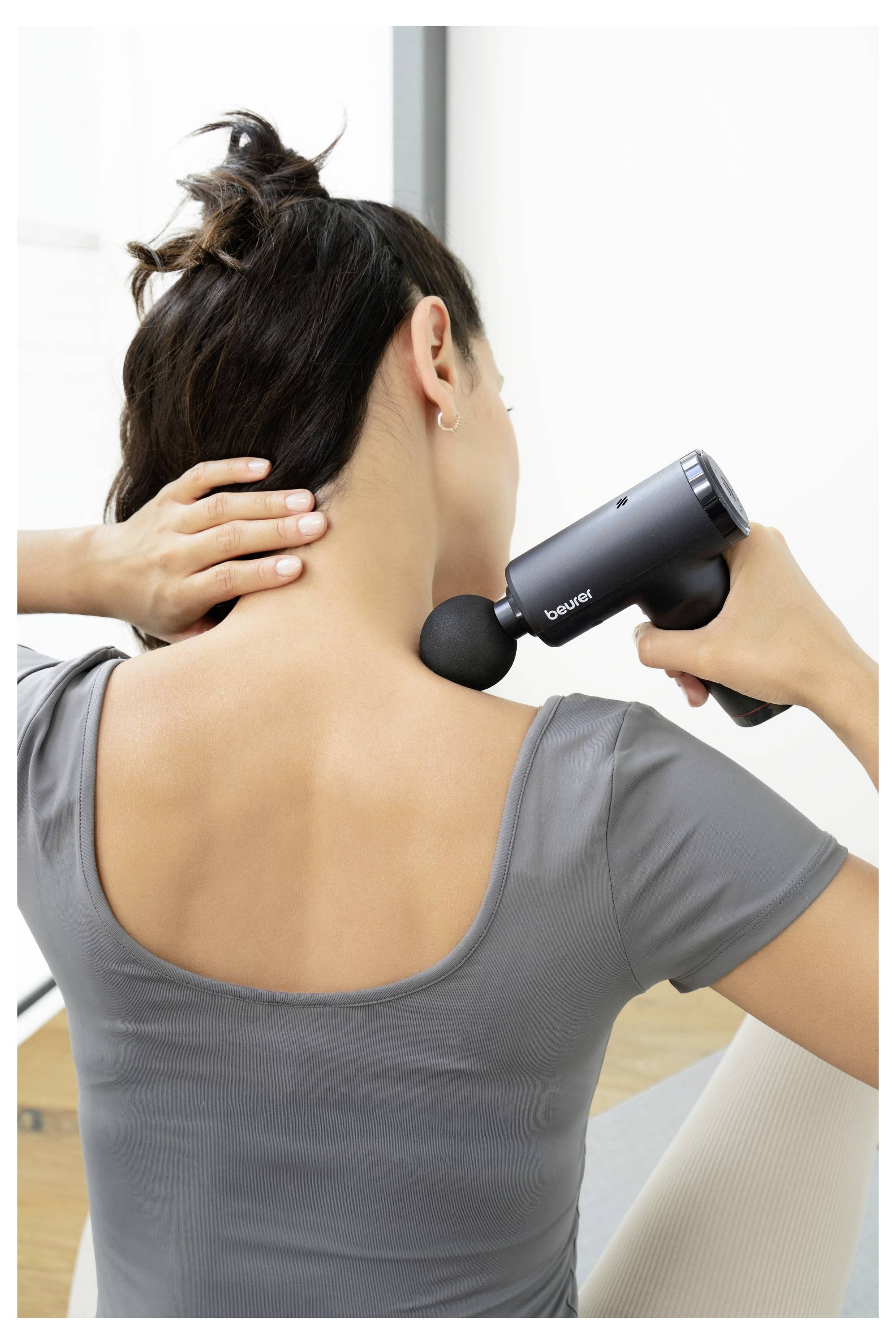 Eine Person nutzt eine schwarze Massagepistole an ihrem Nacken und sorgt so für Muskelentlastung. Sie trägt ein graues Oberteil und sitzt in einem hell erleuchteten Raum.