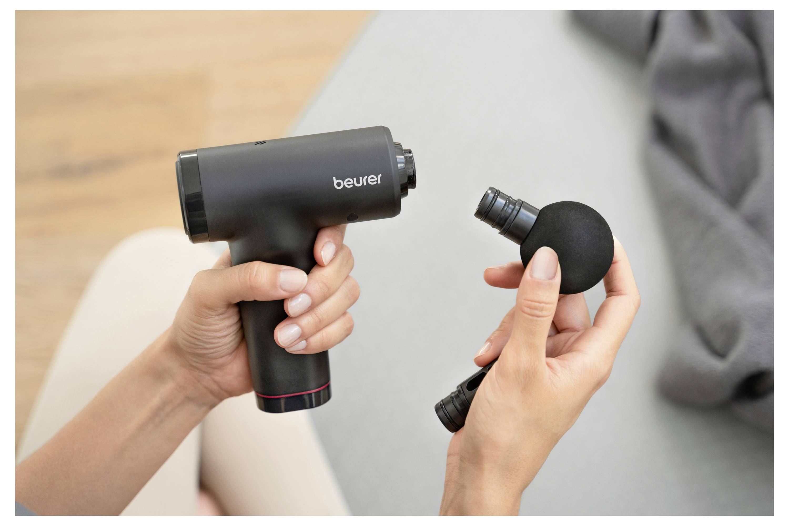 Eine Person hält eine schwarze Massagepistole in einer Hand und befestigt mit der anderen Hand einen kugelförmigen Massagekopf. Ein graues Tuch liegt in der Nähe.