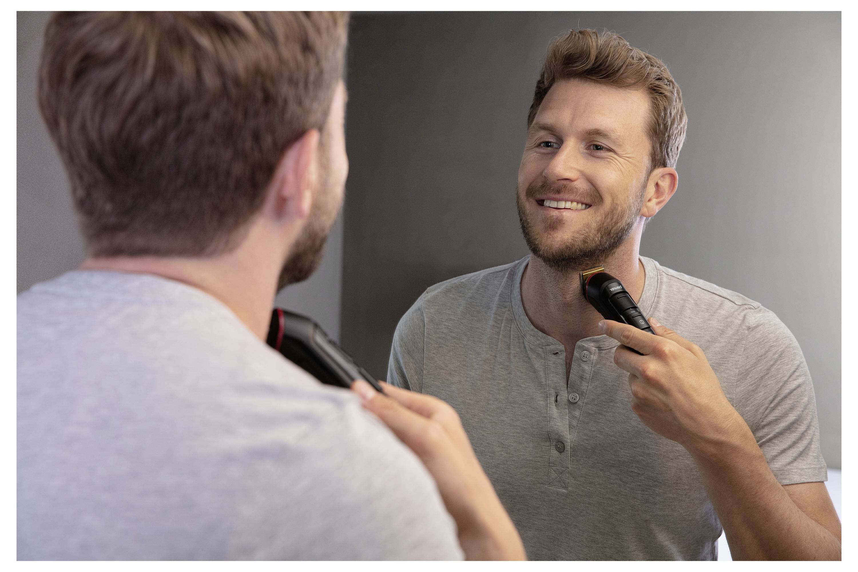 Eine Person steht vor einem Spiegel, lächelt und trimmt sich mit einem elektrischen Rasierer den Bart.