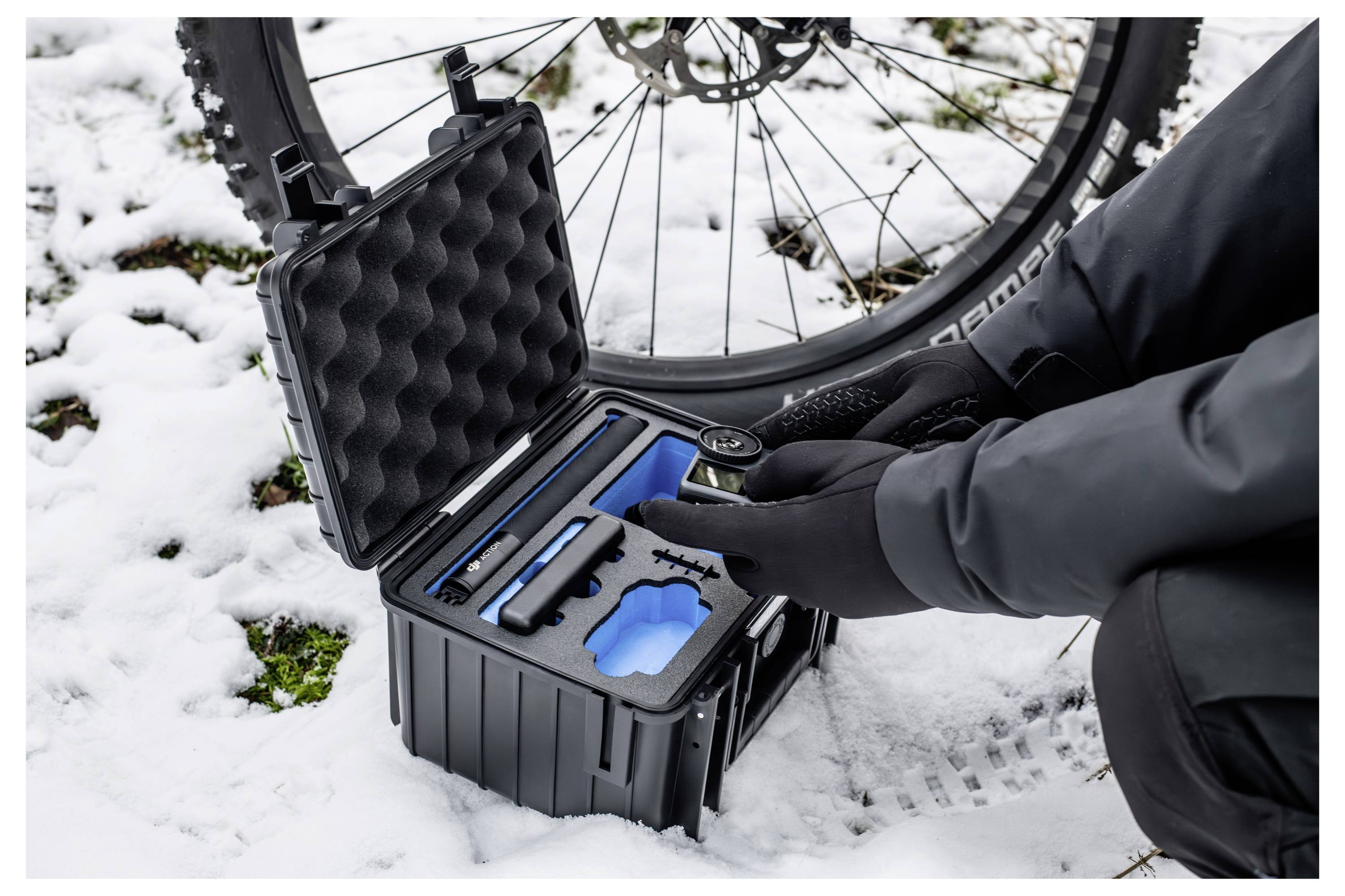 Eine Person in Handschuhen kniet neben einem Fahrradreifen im Schnee und richtet Werkzeuge in einem schwarzen Koffer ein, der für die Fahrradwartung im Freien konzipiert ist.
