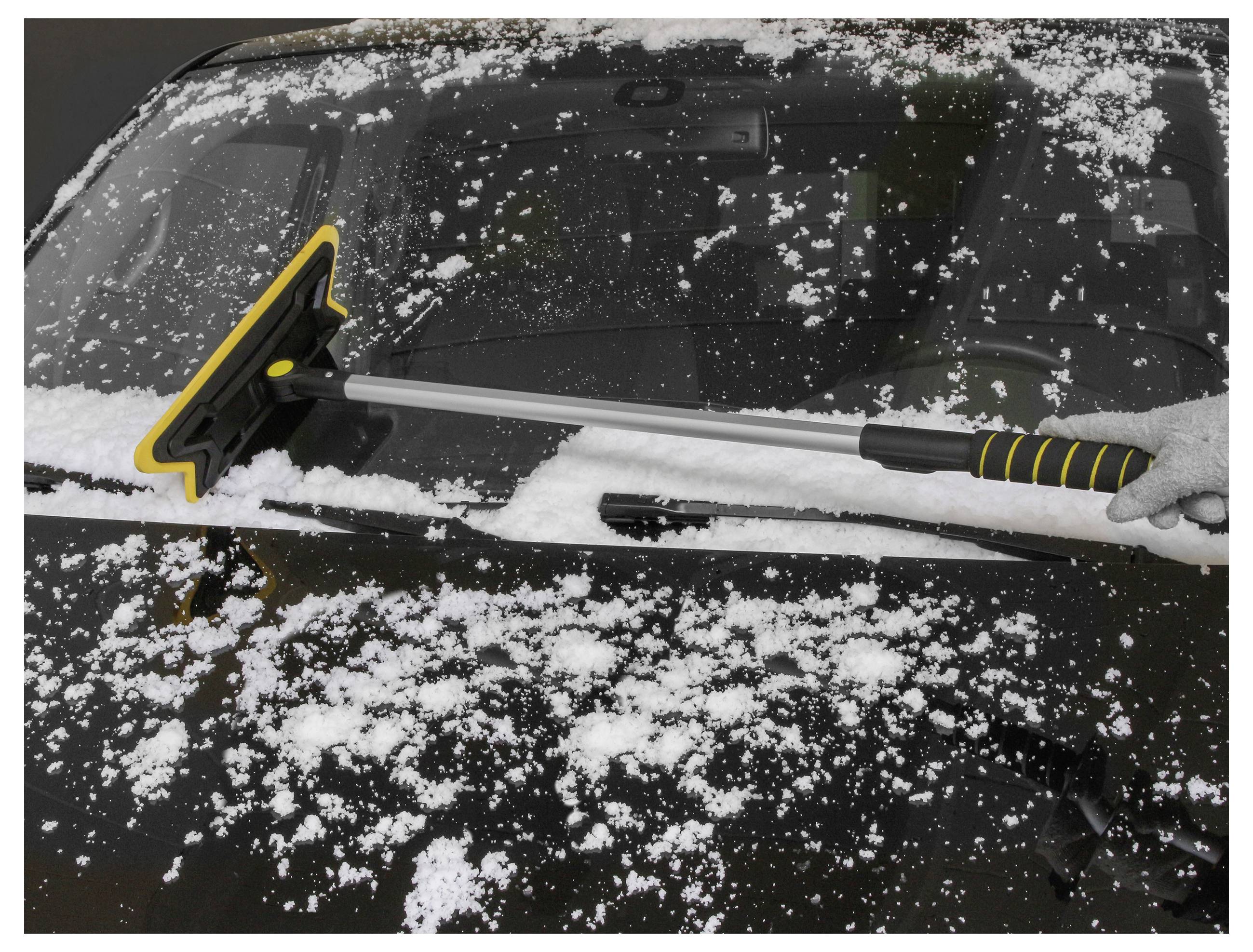 Eine Hand benutzt eine Schneebürste, um Schnee von der Windschutzscheibe eines Autos zu entfernen, was Winterwetter und Fahrzeugwartung demonstriert.