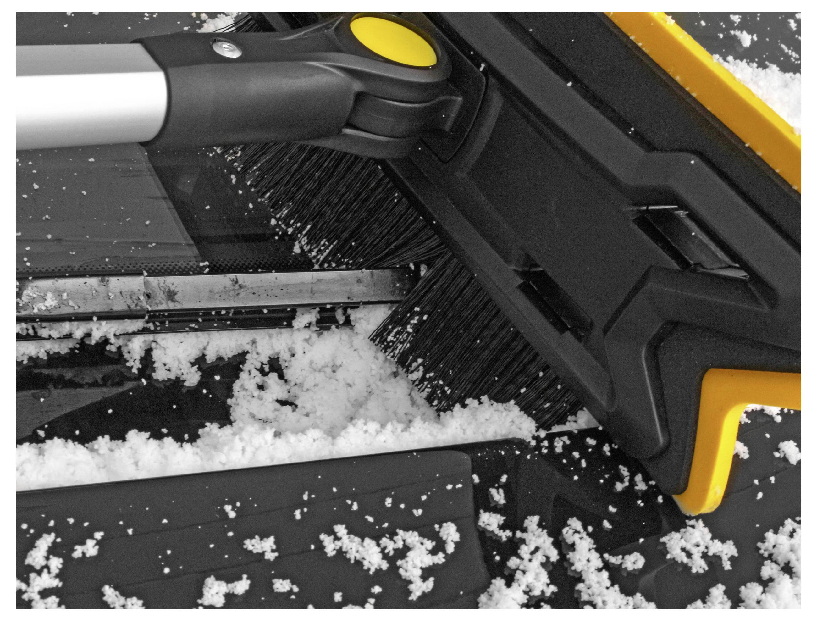 Eine Schneebürste mit gelbem Griff räumt Schnee von der Windschutzscheibe eines Autos. Die Autofläche ist teilweise mit Schneeflocken bedeckt.