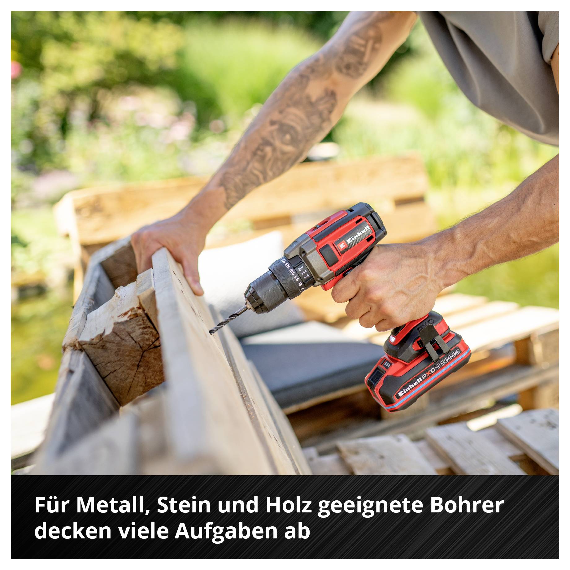 Ein roter Akku-Bohrer wird von einer Person benutzt, um Holz zu bohren. Im Hintergrund sind verschwommene Gartenmöbel zu sehen.
