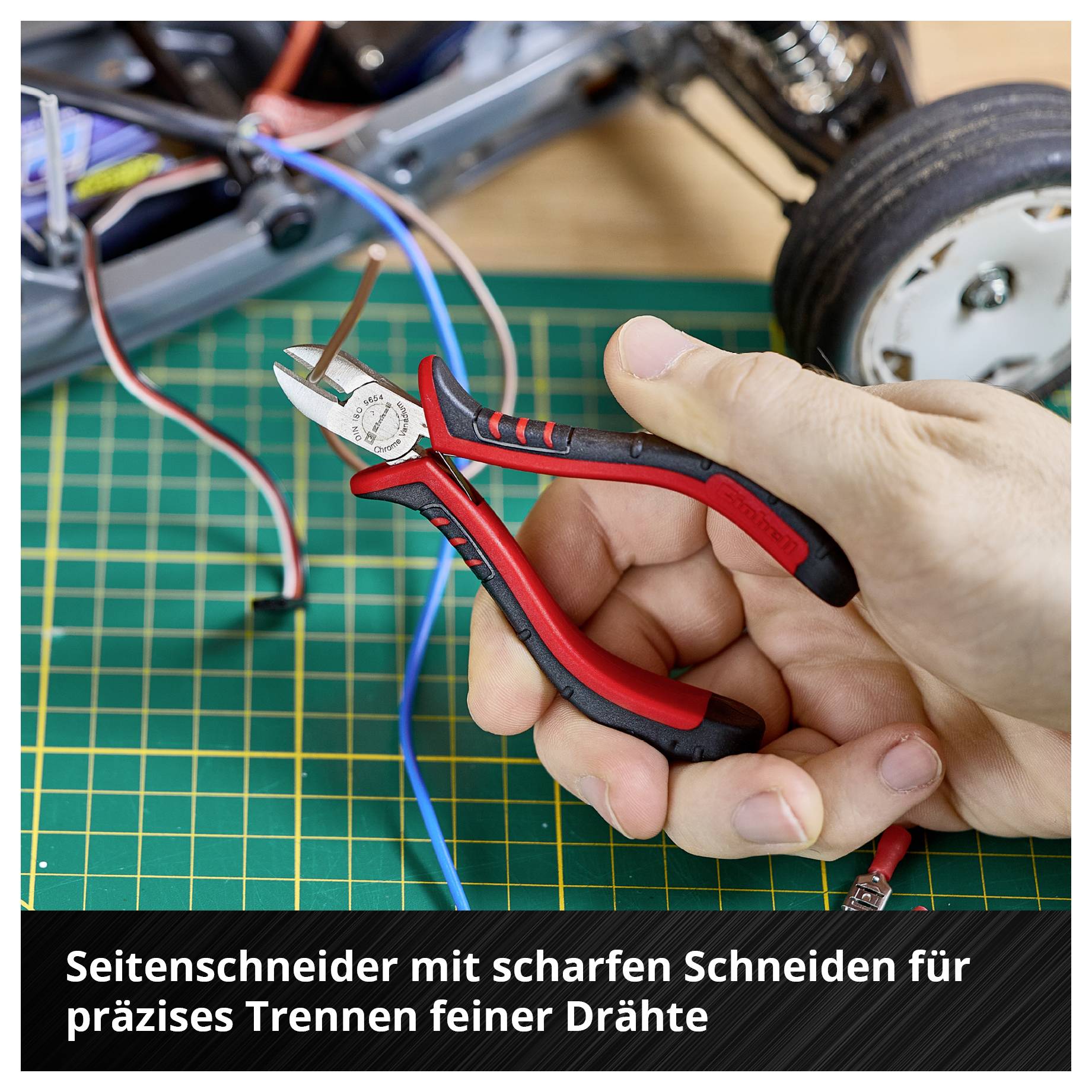Eine Hand hält einen Seitenschneider über einem grünen Schneidebrett. Im Hintergrund sind Drähte und Teile eines Geräts sichtbar.