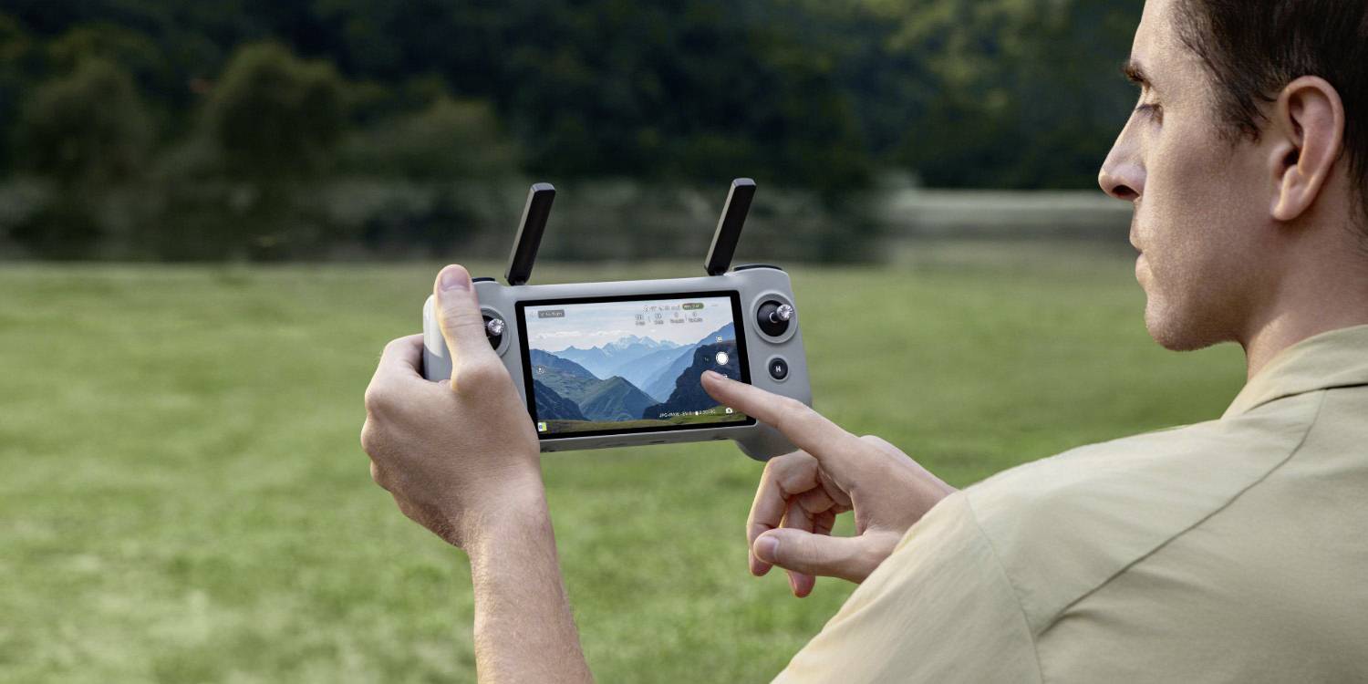 Eine Person, die eine Drohnensteuerung hält, konzentriert sich auf den Bildschirm, der einen Live-Feed einer Berglandschaft anzeigt, was auf die Verwendung im Freien hindeutet.