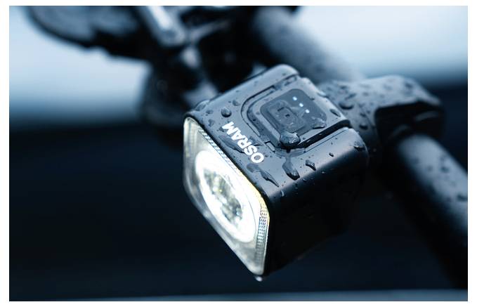 Eine Nahaufnahme eines schwarzen Fahrradvorderlichts mit Wassertropfen, montiert auf dem Lenker. Das Licht leuchtet hell und ist von Osram hergestellt.