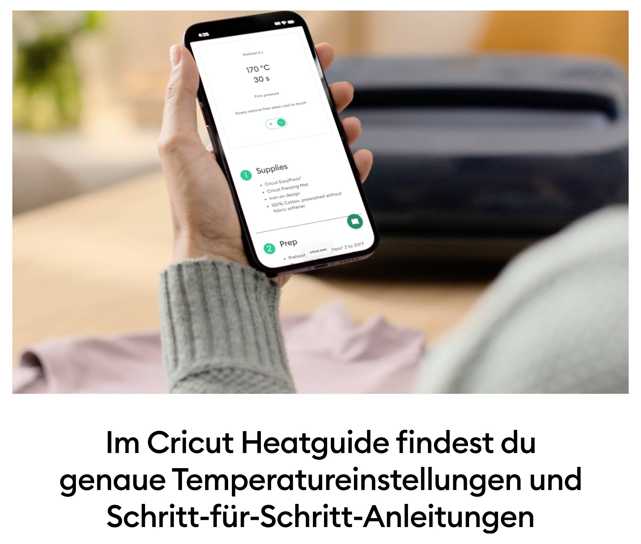 Eine Person hält ein Smartphone mit der Cricut Heatguide-App, die Temperaturen und Anleitungen für Projekte zeigt. Auf einem Tisch liegt ein Cricut-Gerät.