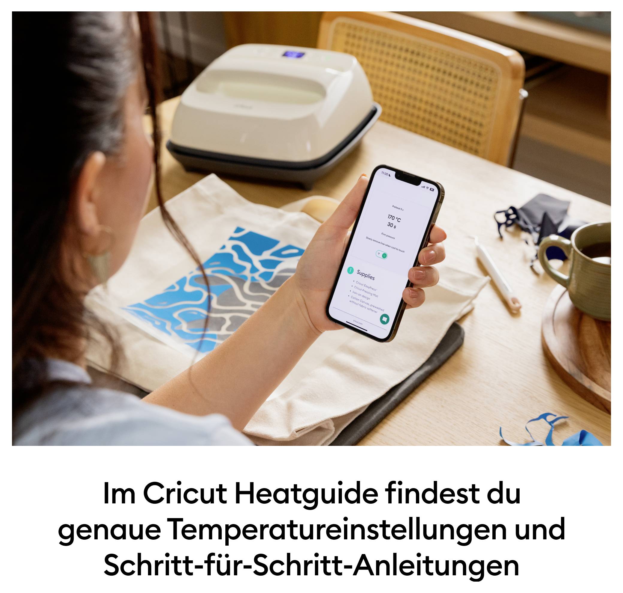 Eine Person hält ein Smartphone mit geöffneter Cricut Heatguide-App. Im Hintergrund sind eine Transferpresse und ein Stoffprojekt zu sehen.