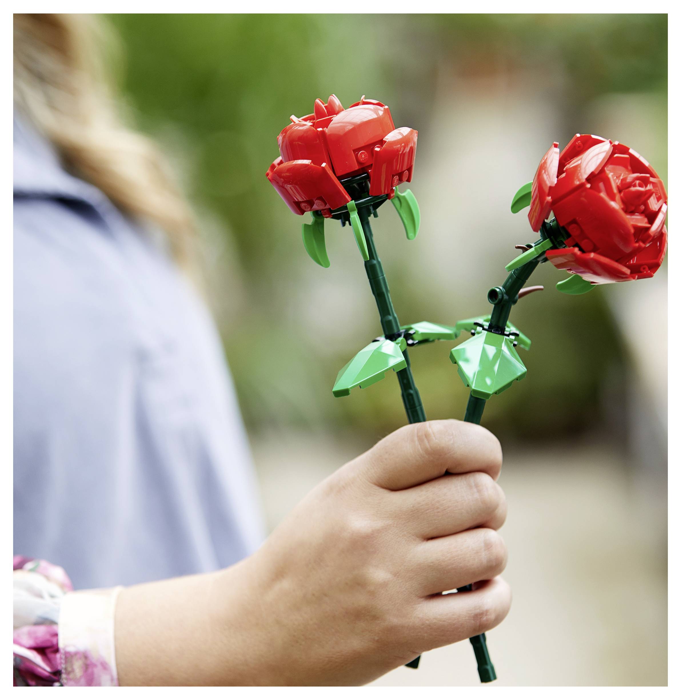 Eine Person hält zwei rote Lego-Rosen, eine in jeder Hand, in einer Außenumgebung.