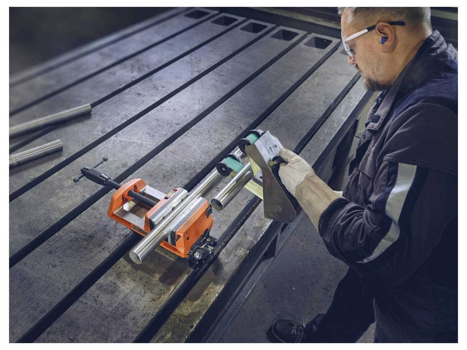 Eine Person bedient ein Schneidewerkzeug an Metallrohren, die in einer orangefarbenen Spannvorrichtung auf einem großen industriellen Werkstatt­tisch gesichert sind. Schutzausrüstung wird getragen.