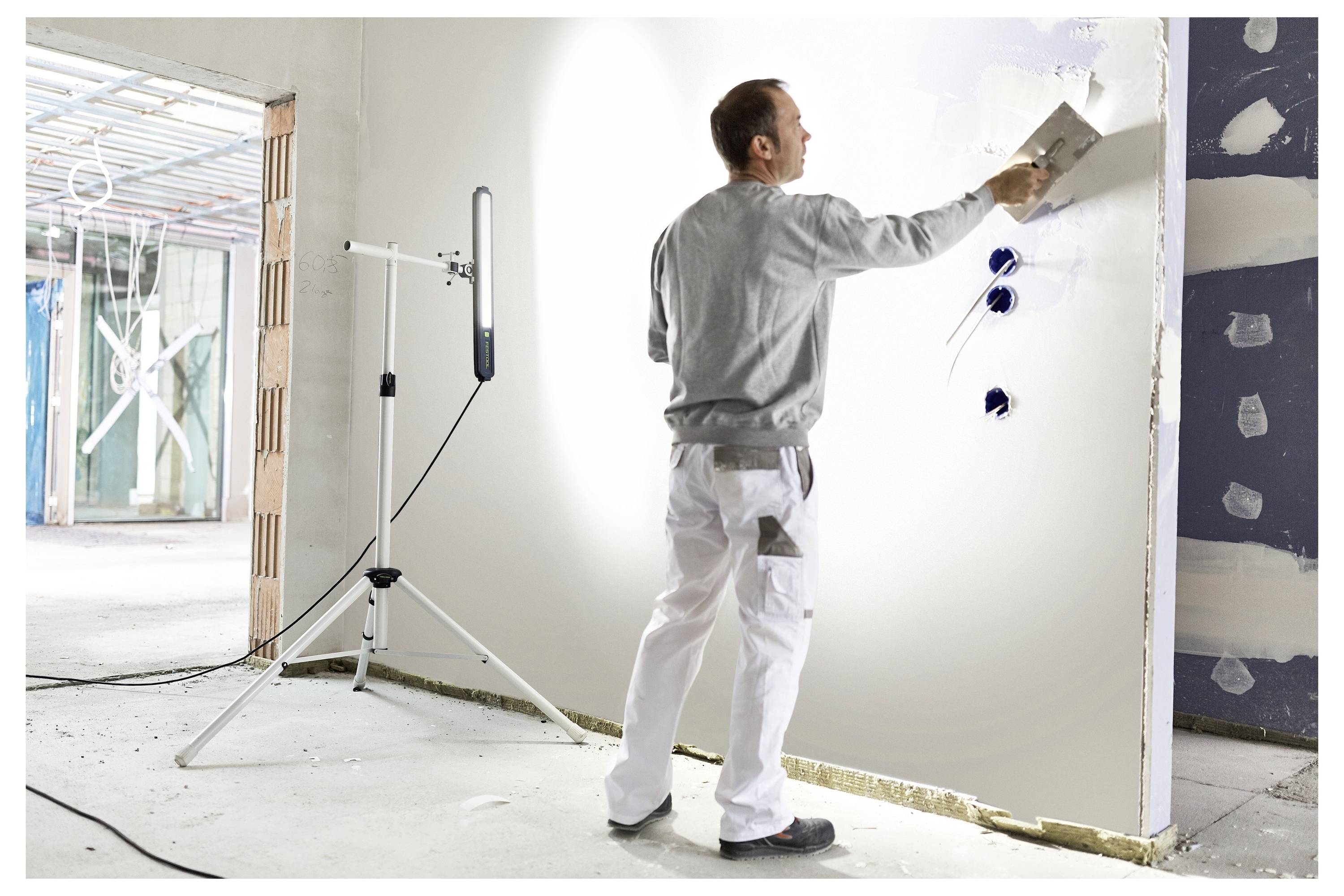 Ein Arbeiter in weißer Hose und grauem Hemd glättet mit einer Kelle Gips auf einer teilweise konstruierten Wand in einem Gebäude im Bau.